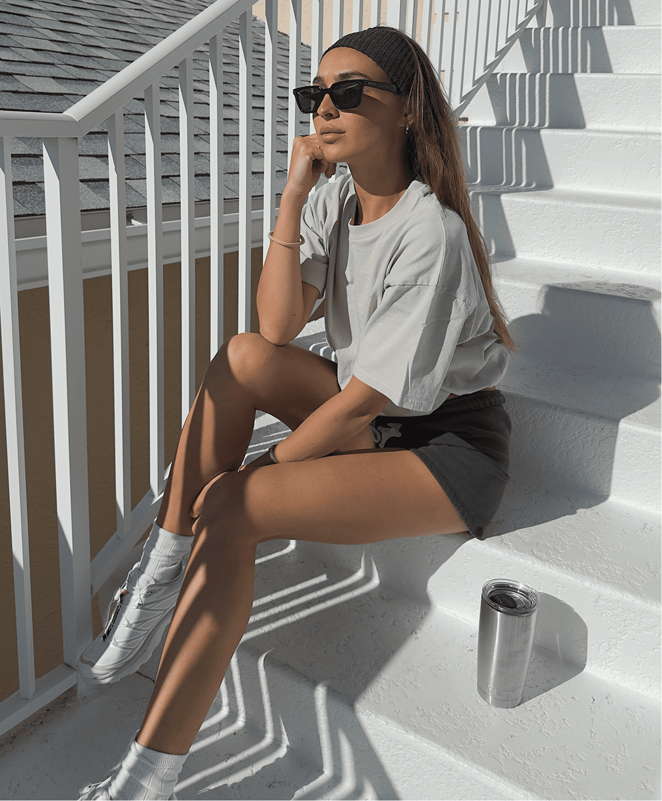 Woman sitting on white outdoor steps, wearing sunglasses, black headband, oversized gray T-shirt, black shorts, white sneakers, with a stainless steel travel mug nearby.