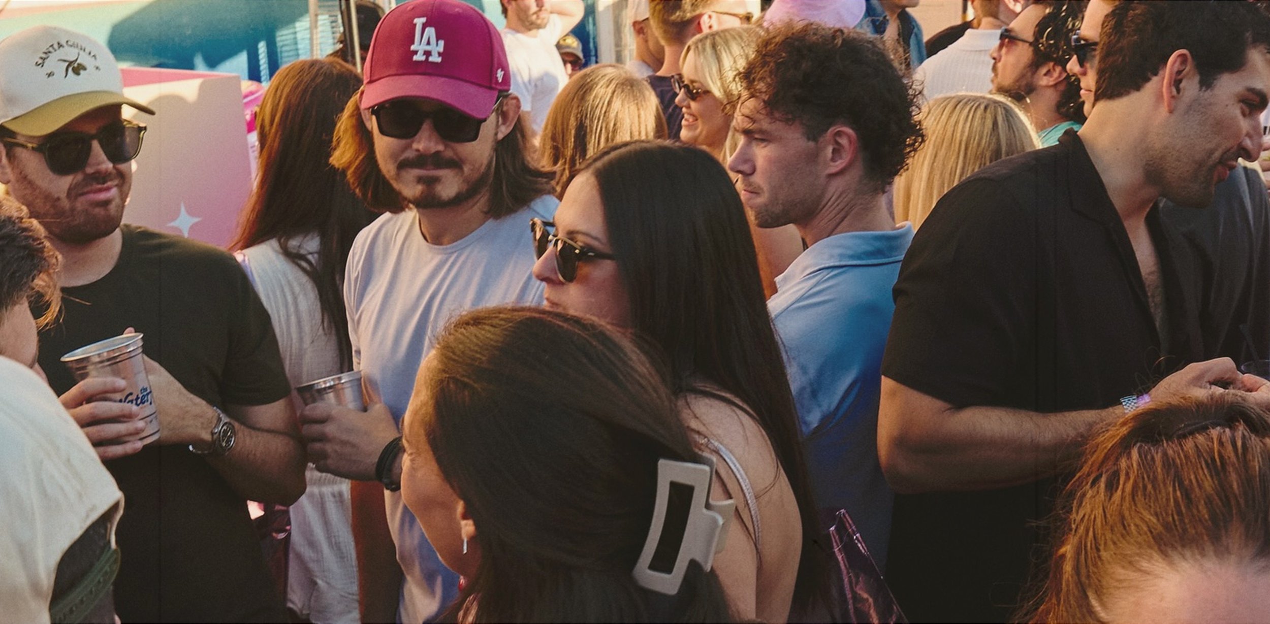 A crowd of people at an outdoor event, many wearing sunglasses and holding drinks, with a mix of ages and ethnicities, suggesting a social gathering or festival.