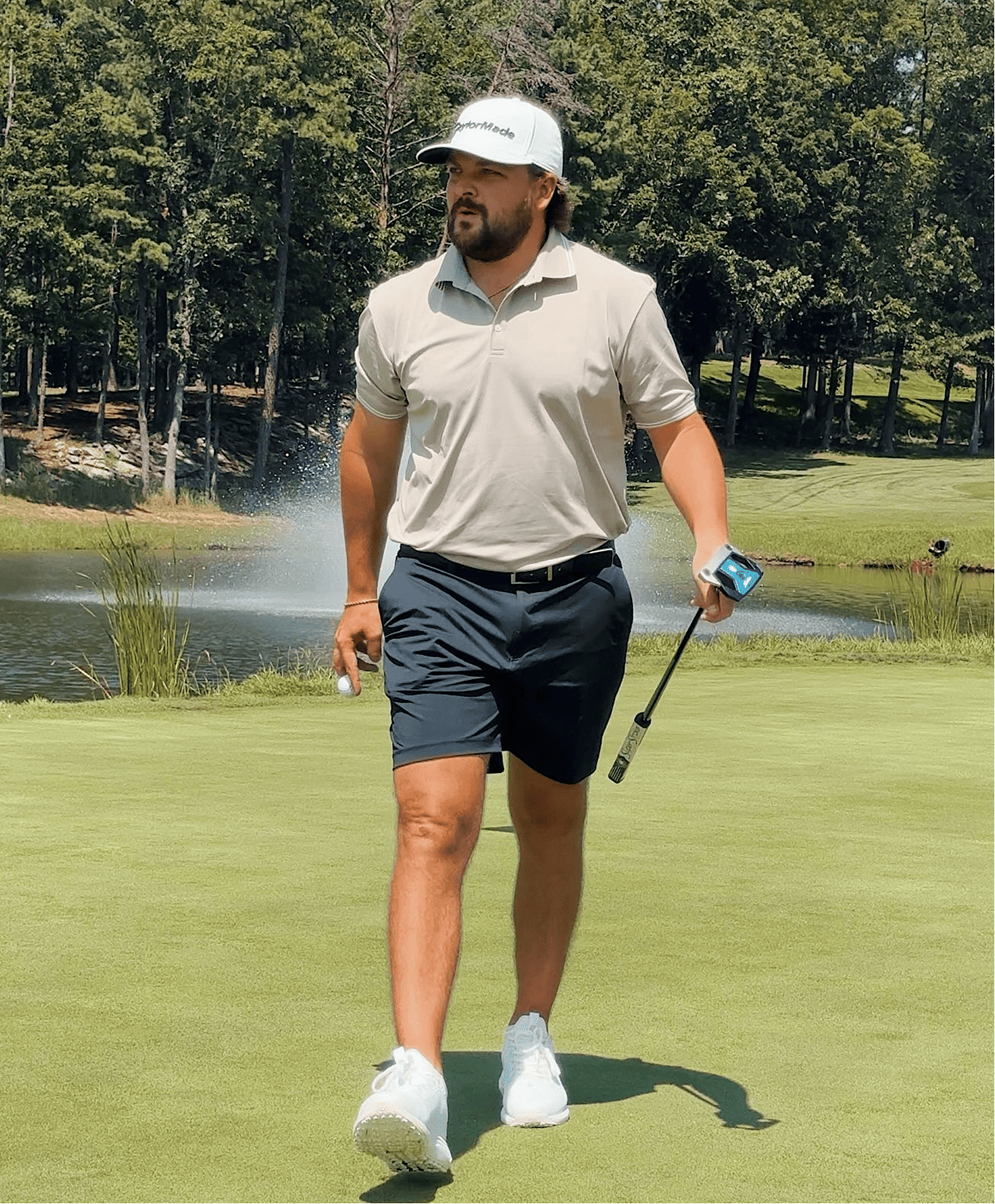 A man walking on a golf course holding a golf club and a golf ball, with a small lake and trees in the background.