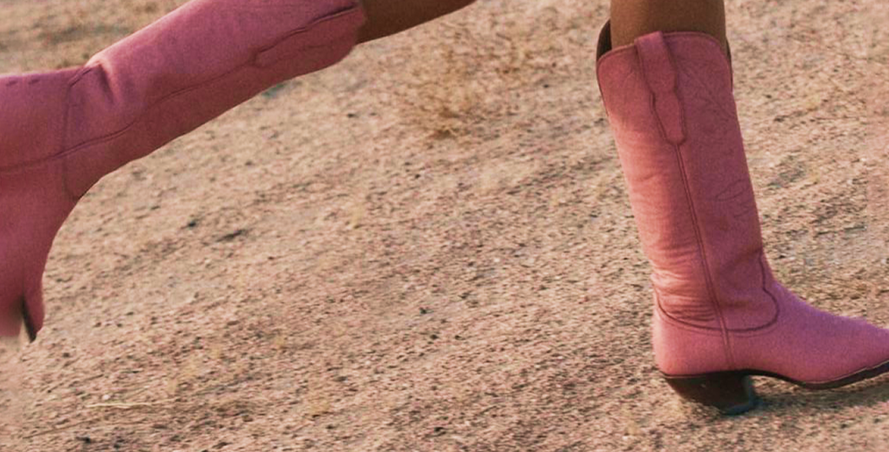 Close-up of a person wearing pink cowboy boots on a sandy surface.