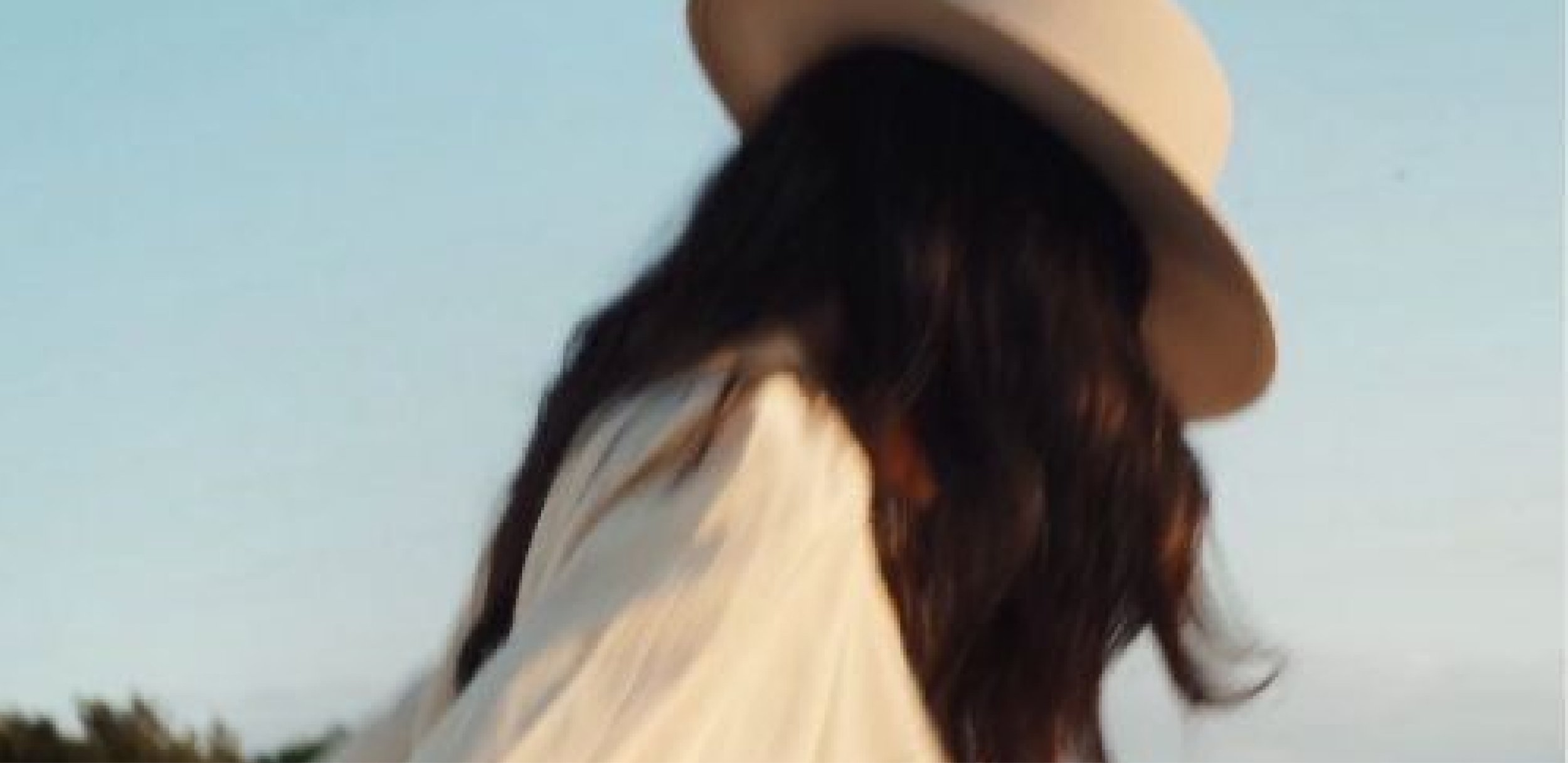 A woman wearing a beige hat with a wide brim and a beige coat, with long dark hair, looking down against a clear sky background.
