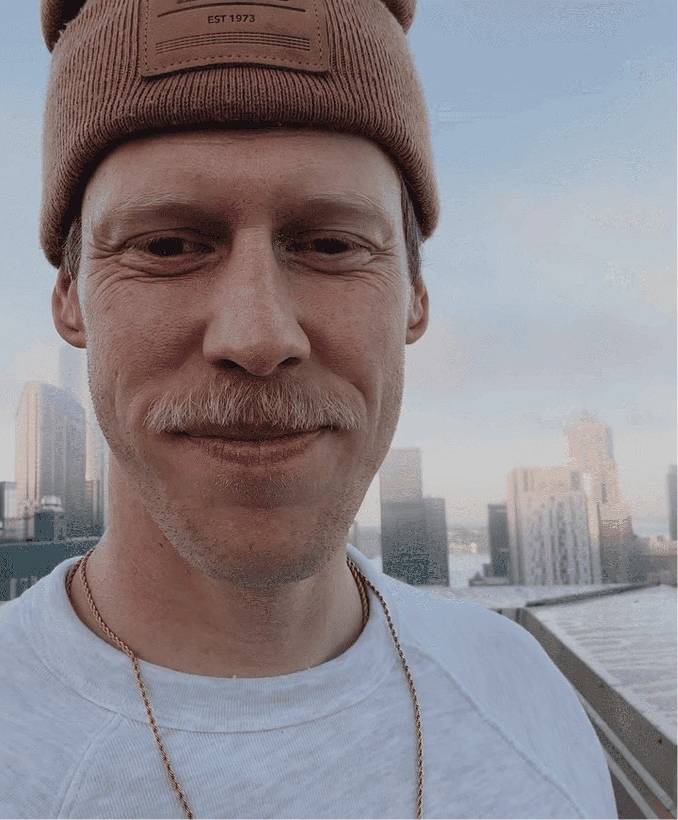 Close-up of a man with a light complexion, light brown mustache, and short hair, wearing a brown beanie and a white t-shirt, standing on a rooftop with a city skyline in the background during daytime.