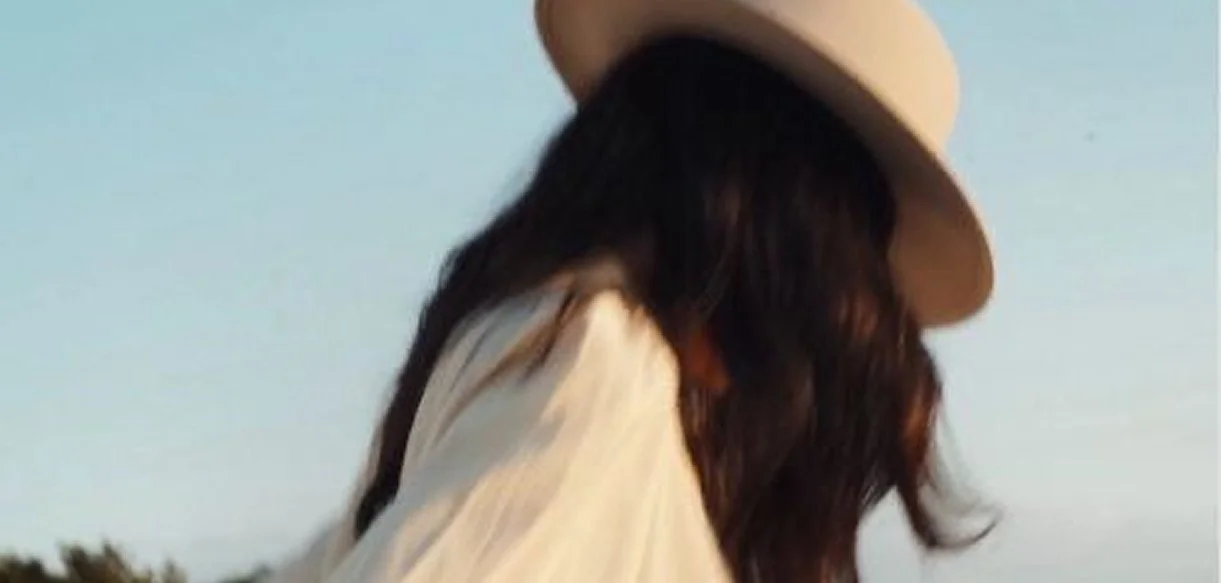 A woman with long dark hair wearing a beige hat, white dress, and looking down outdoors.