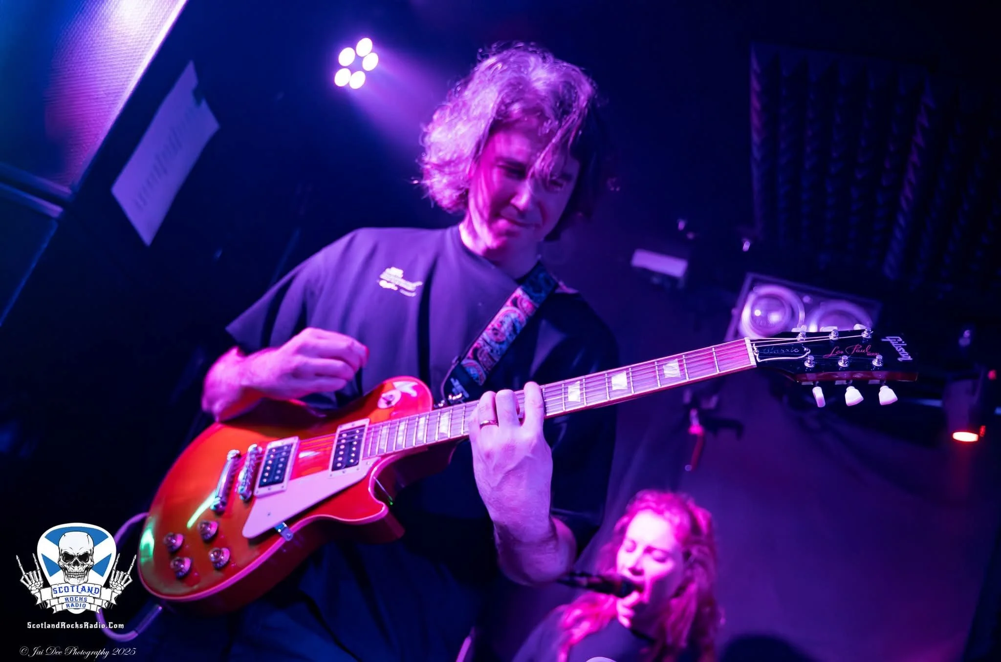 SOMB Scared of My Brother Guitar Player Andy Russell. A person with curly hair playing an orange electric guitar on stage, with purple lighting and SOMB Scared of My Brother Keyboard player Phoebe Russell in the background.