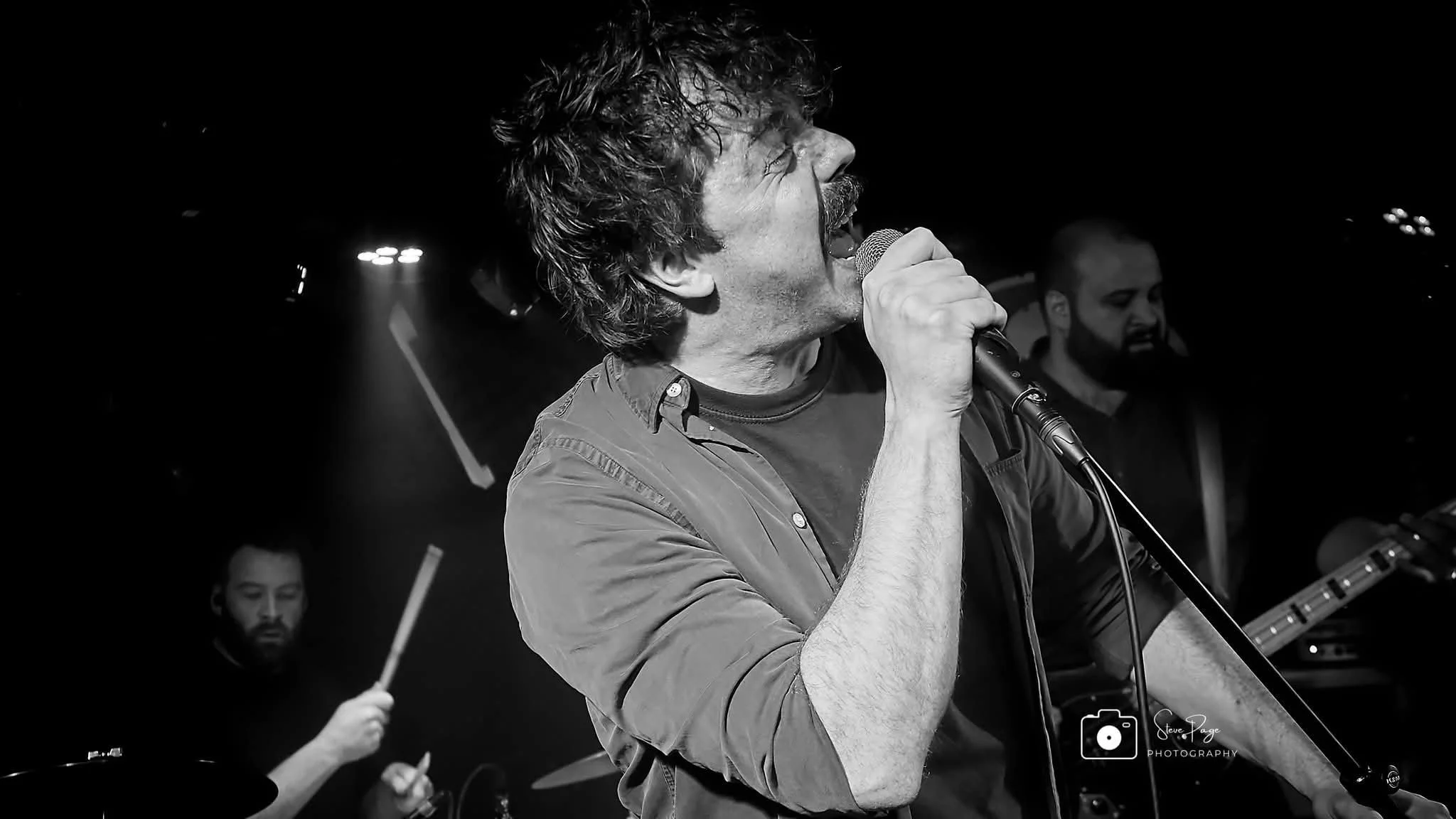 SOMB Scared of My Brother lead singer (Lewis Thomson) singing into a microphone, with two band members playing instruments in the background, on a dark stage with stage lighting.