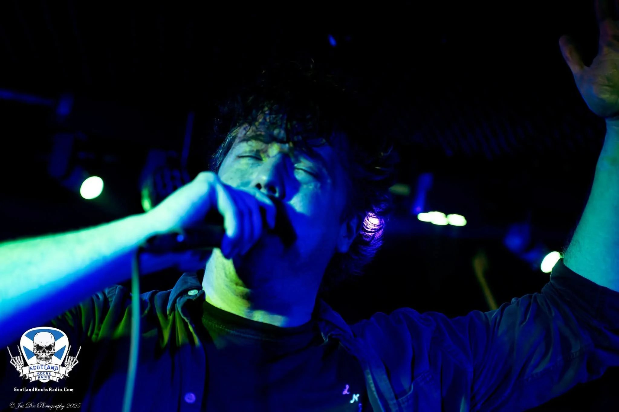 SOMB Scared of My Brother Lead Singer (Lewis Thomson) performing passionately with eyes closed, holding a microphone in a dark, colorful concert setting.