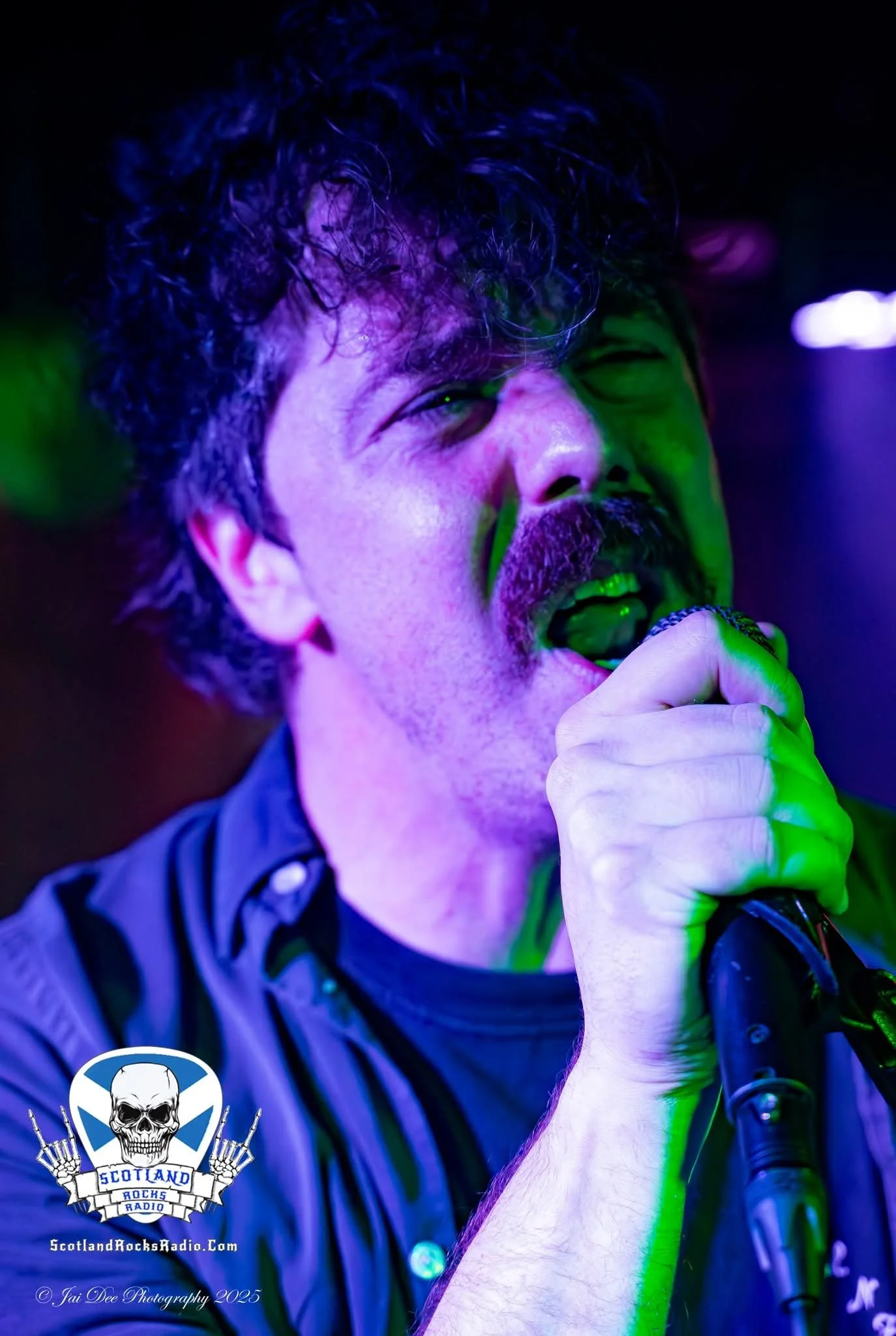SOMB Scared of My Brother lead singer (Lewis Thomson) , illuminated by purple and green stage lights, with a logo for Scotland Rocks Radio in the bottom left corner.