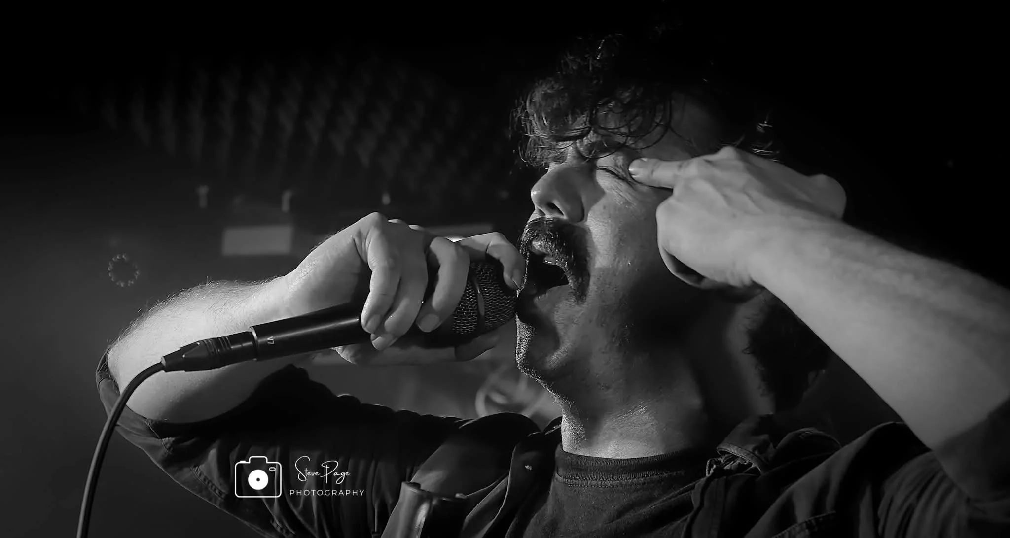 SOMB Scared of My Brother Lead Singer (Lewis Thomson) passionately performs into a microphone, holding his head with both hands, with a blurred background.