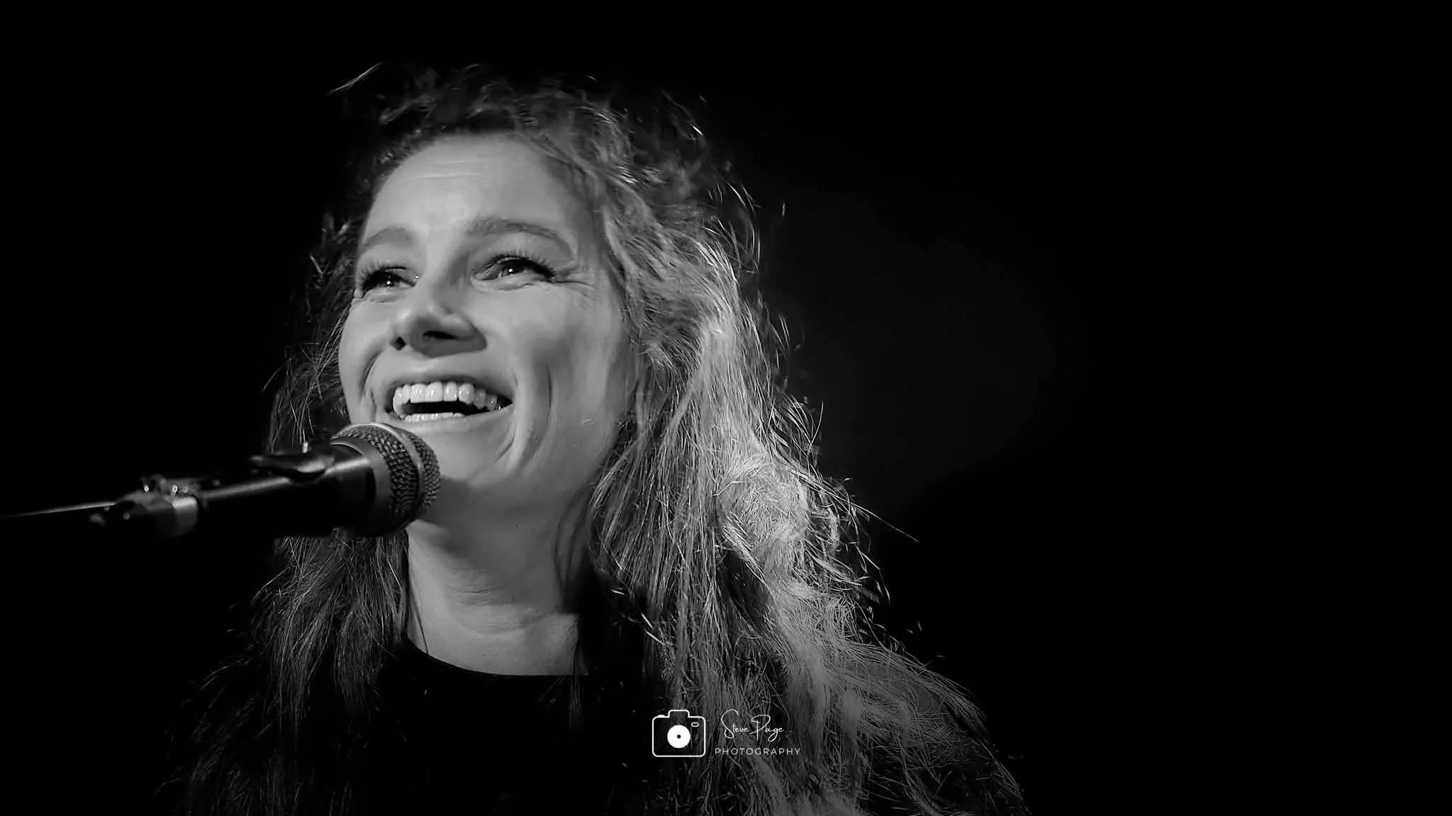 SOMB Scared of My Brother Keyboard player Phoebe Russell  singing into a microphone against a dark background.
