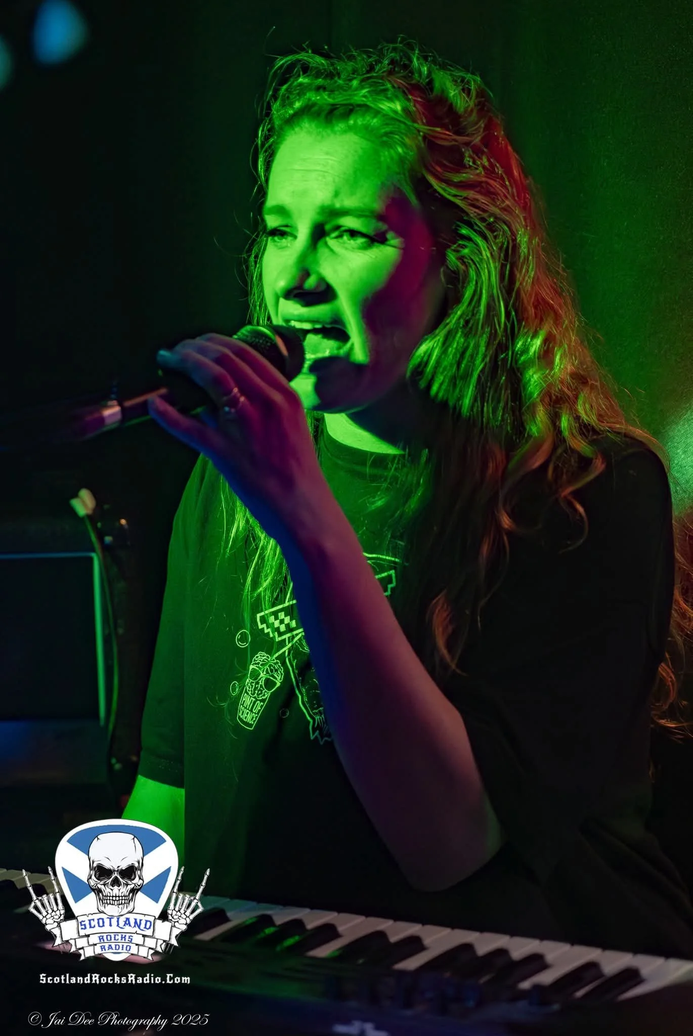 SOMB Scared of My Brother Keyboard player Phoebe Russell  singing into a microphone with green and purple lighting, sitting in front of a piano keyboard, with a logo for Scotland Rocks Radio at the bottom.