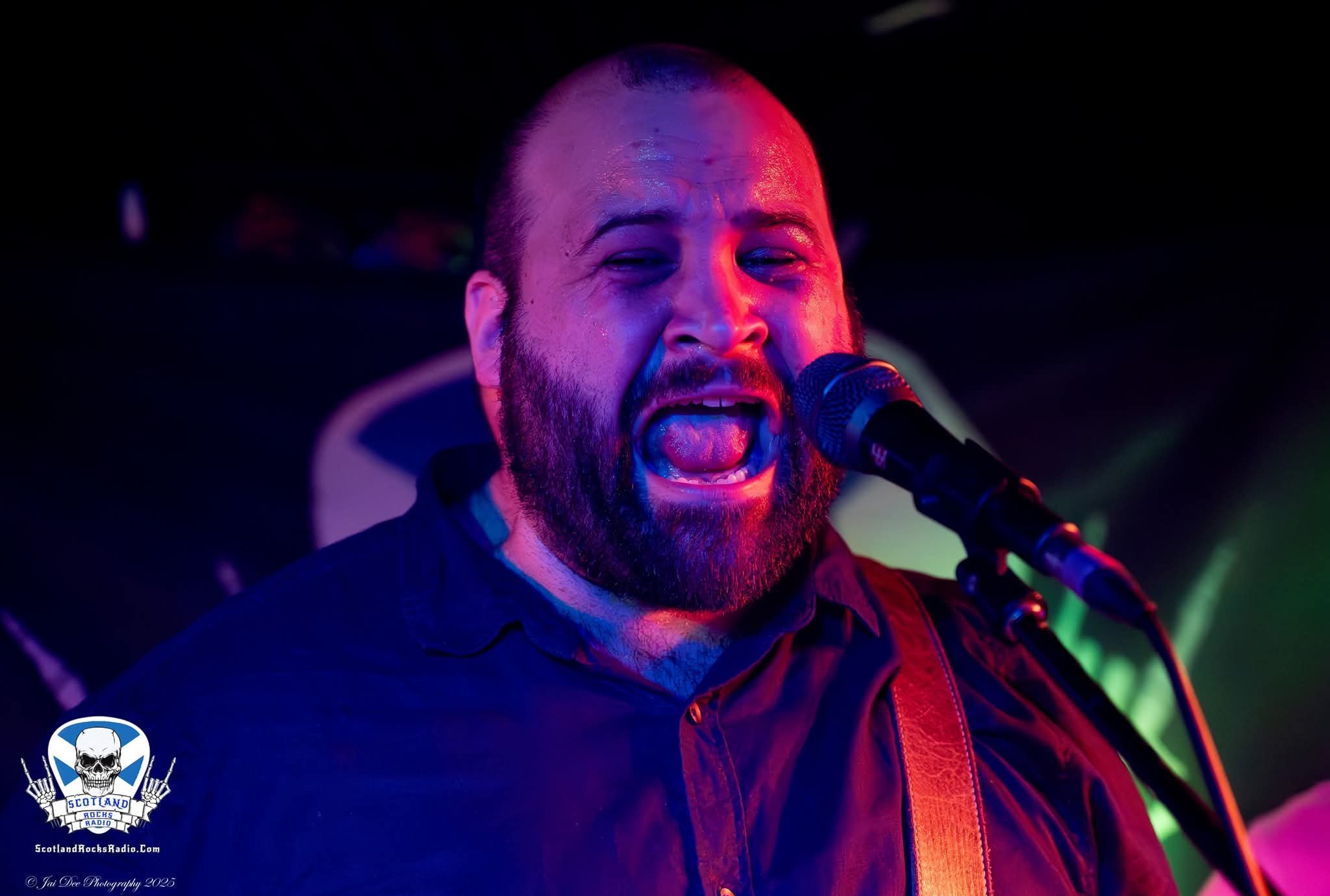 SOMB Scared of My Brother Bass player Toby Kirkwood singing passionately into a microphone with colorful stage lighting in the background.