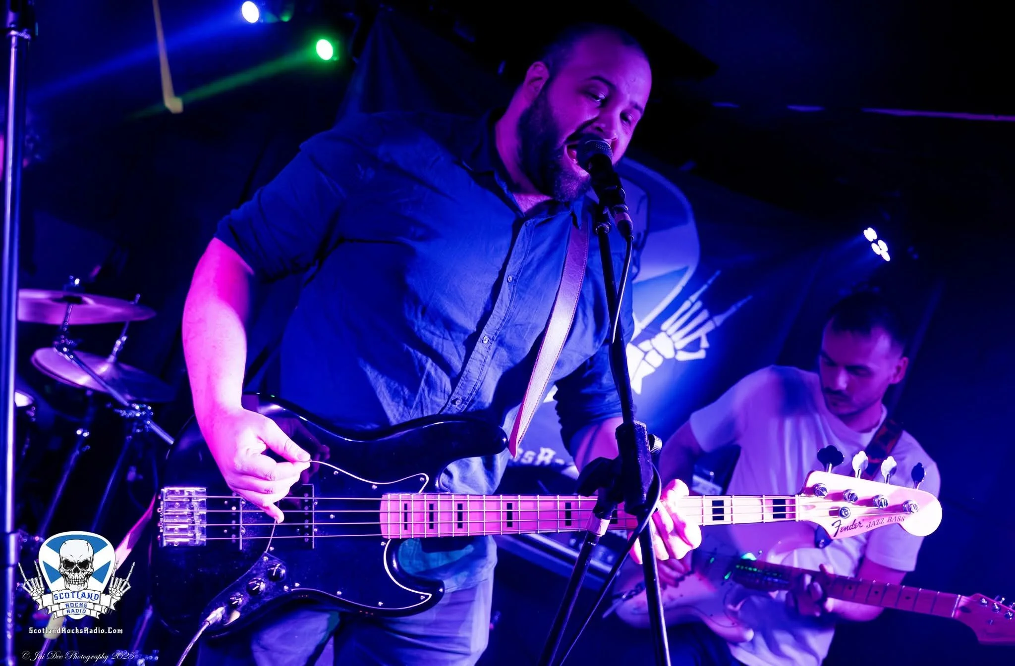 SOMB Scared of My Brother live on stage with Bass Player Toby Kirkwood singing into a microphone and playing an electric bass guitar, and another playing an electric guitar, with stage lighting and a dark background