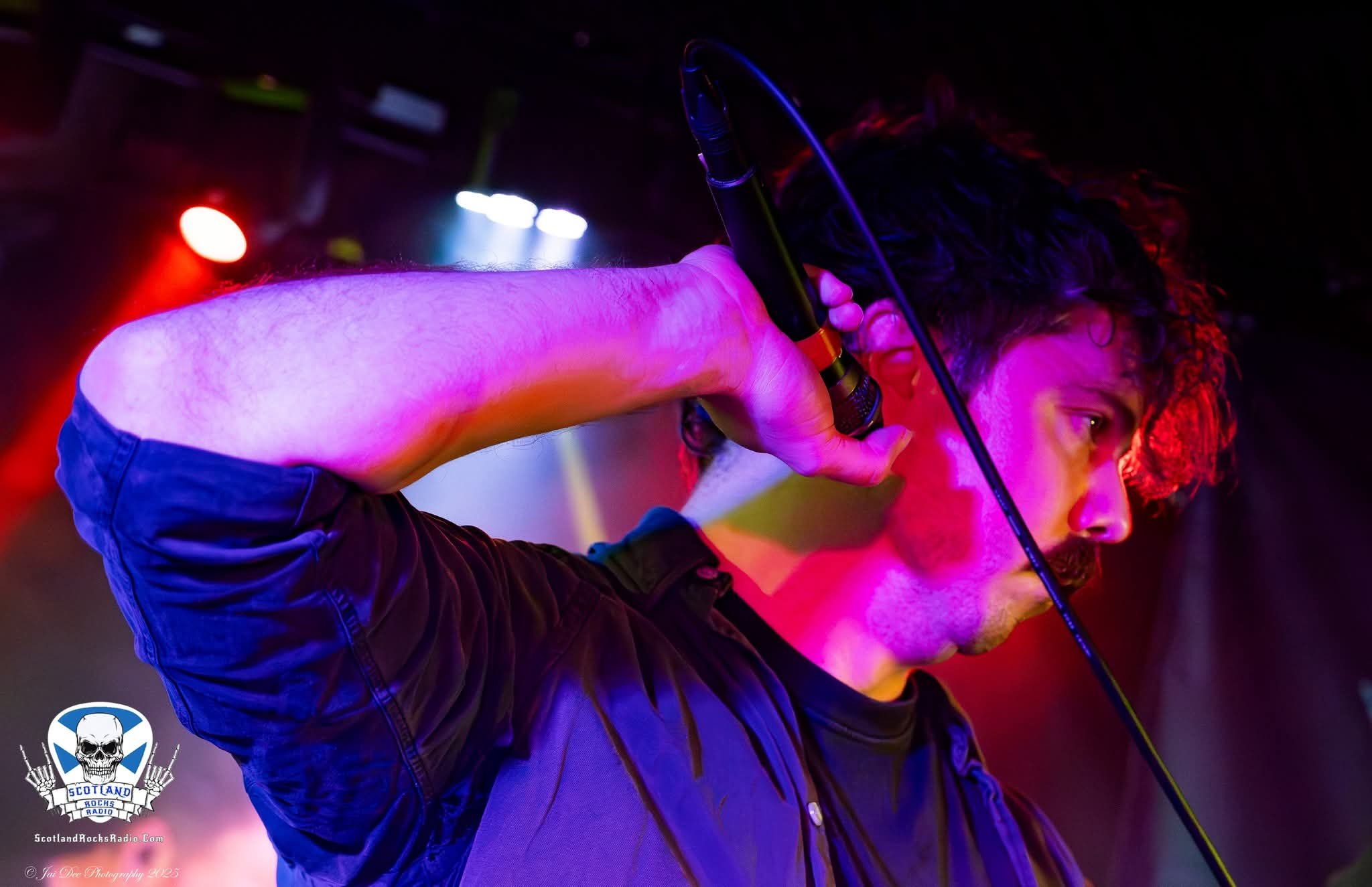 SOMB Scared of My Brother lead singer (Lewis Thomson), singing into a microphone on stage illuminated by pink and purple stage lights.