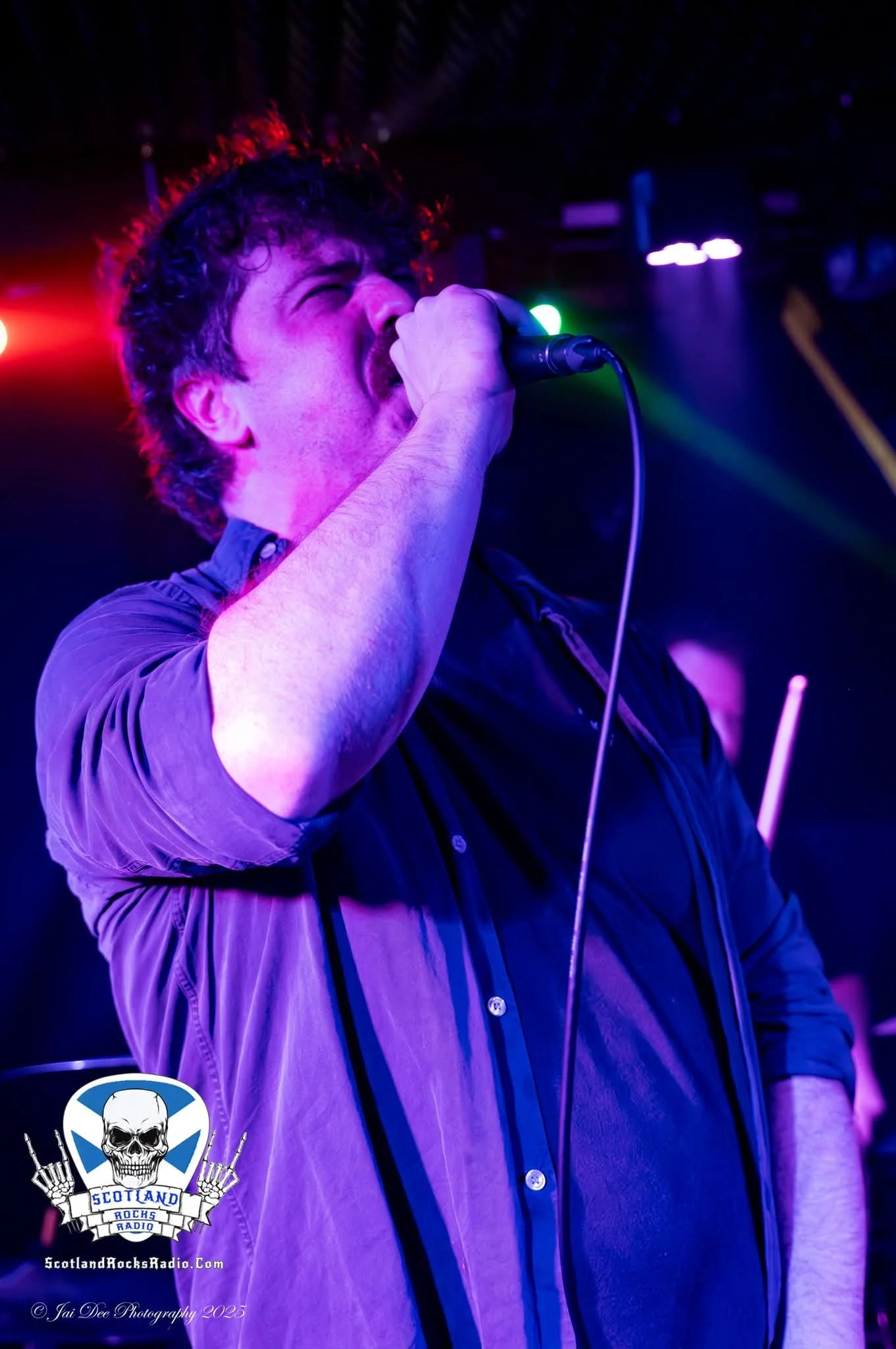A male singer passionately performing on stage with colorful lighting, holding a microphone close to his mouth, wearing a dark shirt with rolled-up sleeves.