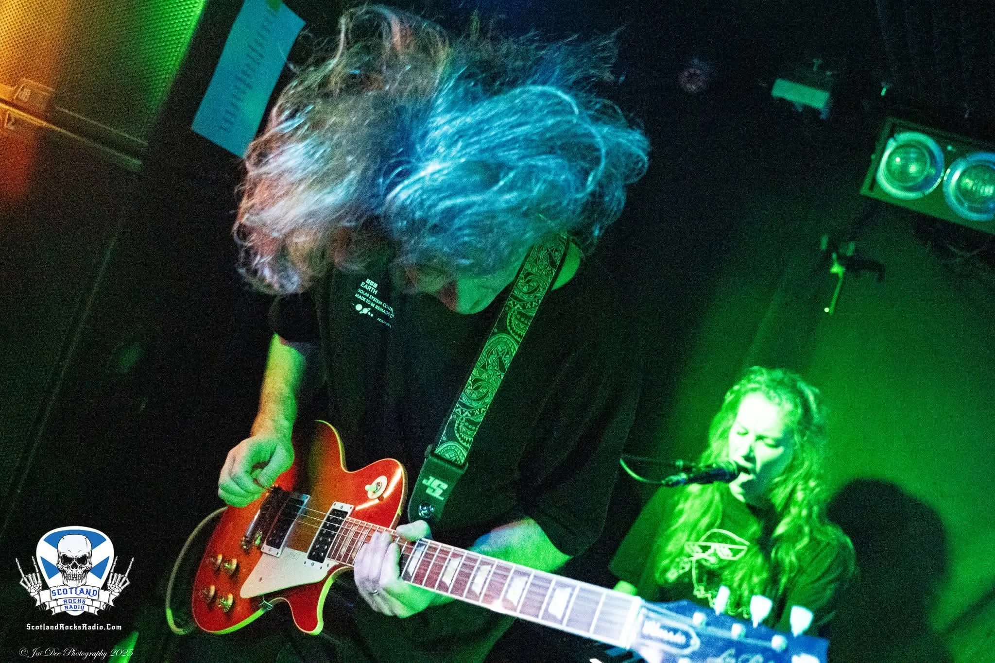 SOMB Scared of My Brother guitar player (Andy Russell) playing a red electric guitar on stage, with keyboard player (Phoebe Russell) singing into a microphone nearby, both illuminated by green stage lights.