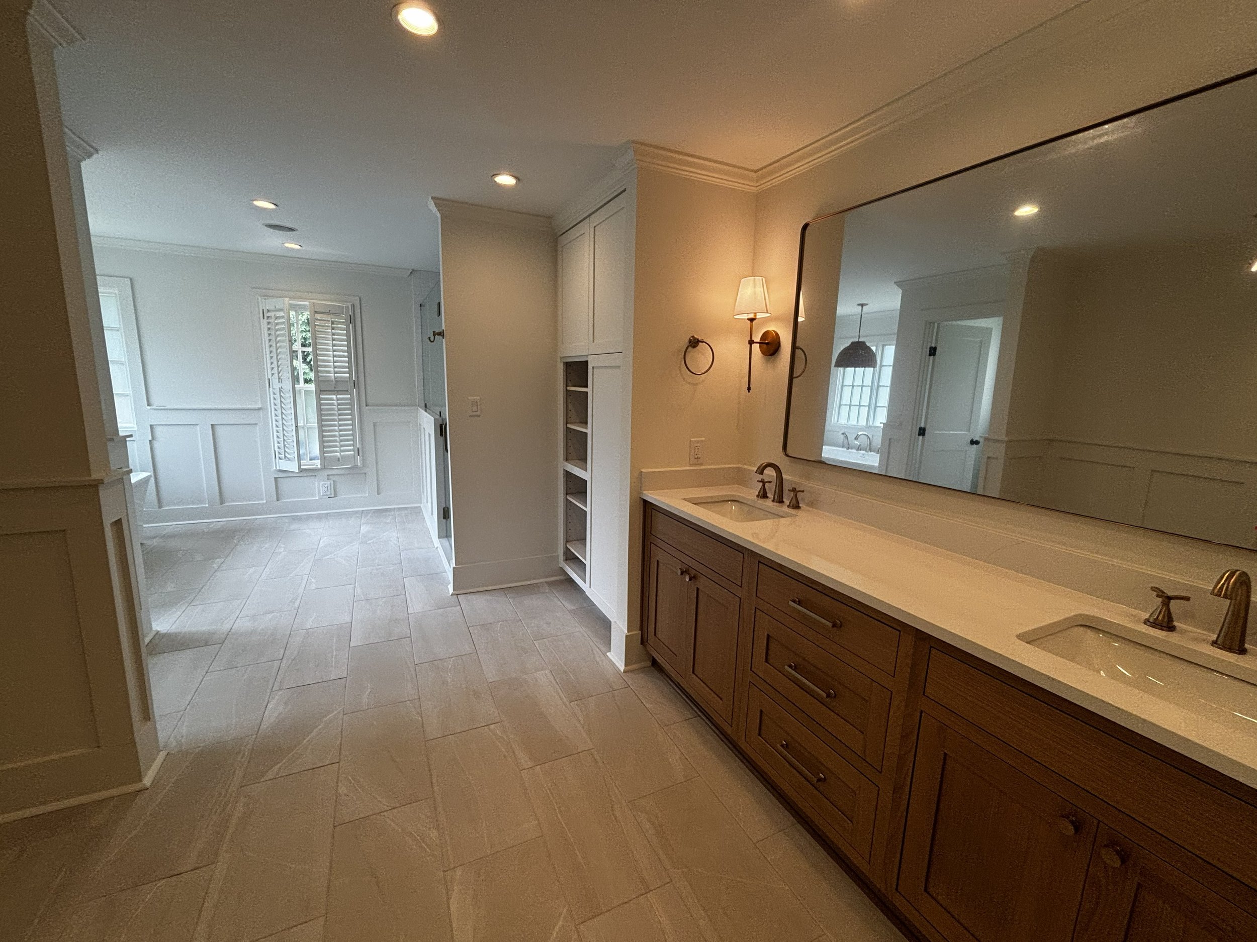 Bathroom with beige tile flooring, white walls, a large mirror above a wooden vanity with a light-colored countertop and a sink, and a wall-mounted light fixture. There is a laundry area or secondary room visible in the background with a window and w