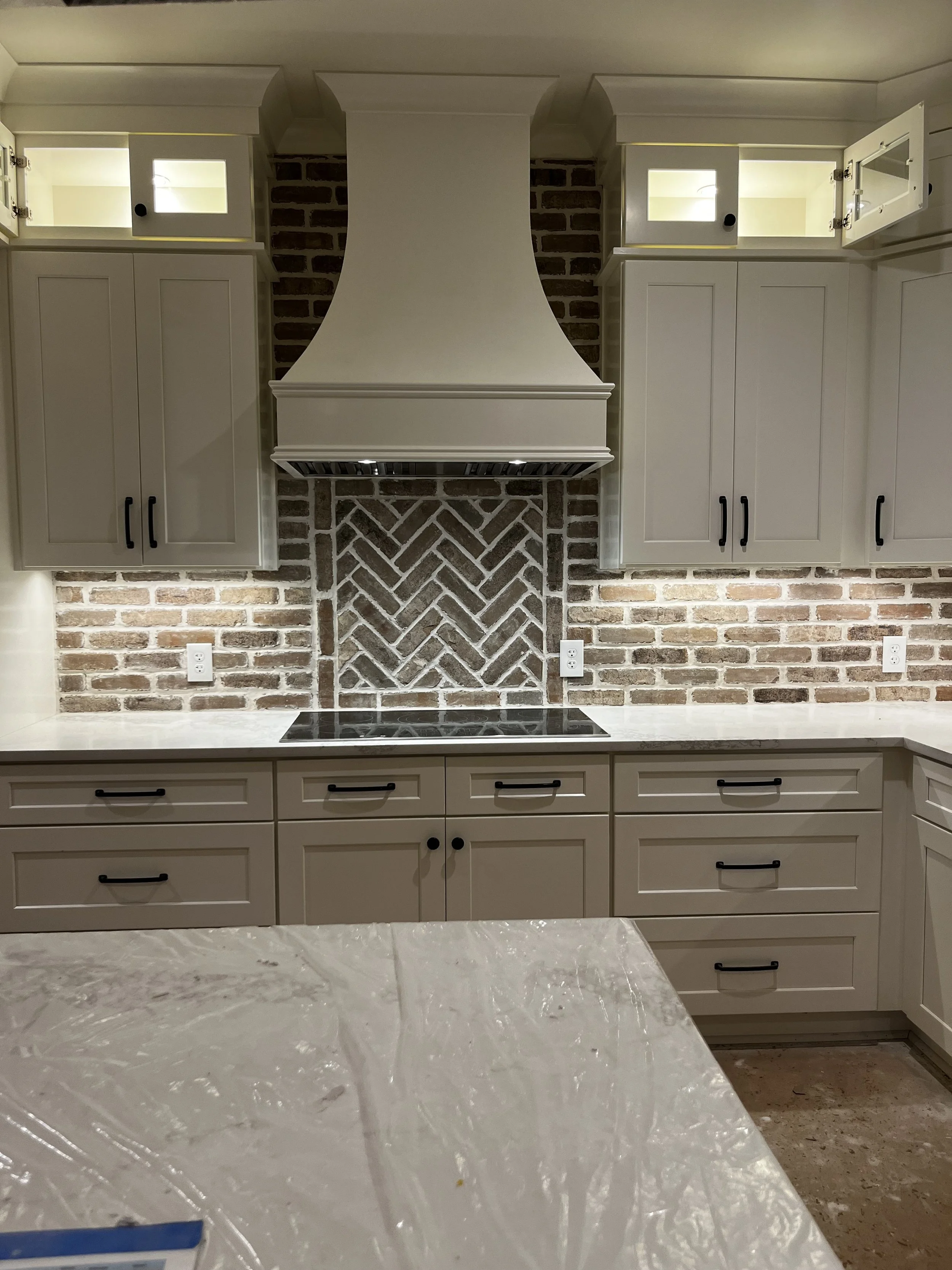 Kitchen with white cabinets, brick wall backsplash, and a white range hood over a black stovetop. Upper cabinets have glass doors with interior lighting. Countertop is white marble.
