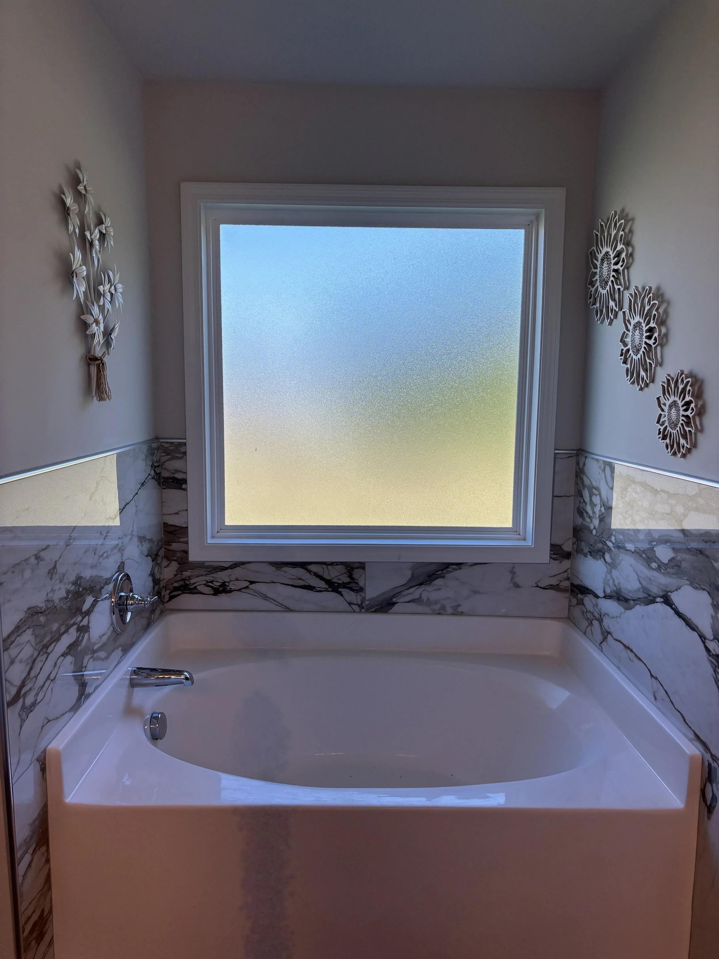 A white bathtub with silver fixtures and marble tiling, situated under a frosted window with decorative wall art on either side.