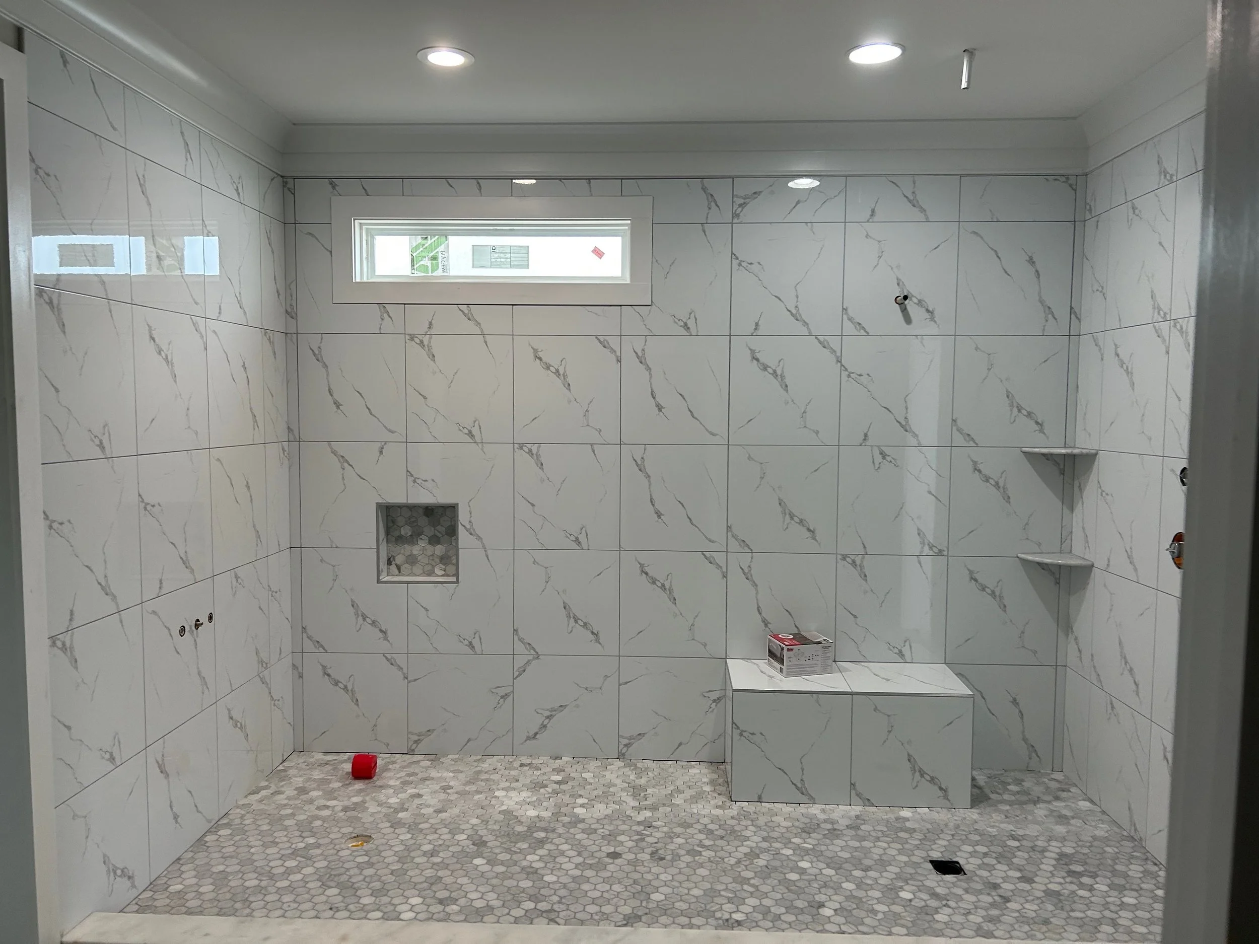A bathroom with white marble tiles with gray veining on the walls, a small rectangular window, and a mosaic tile floor. There are no fixtures installed yet, except for some plumbing poking through the wall.