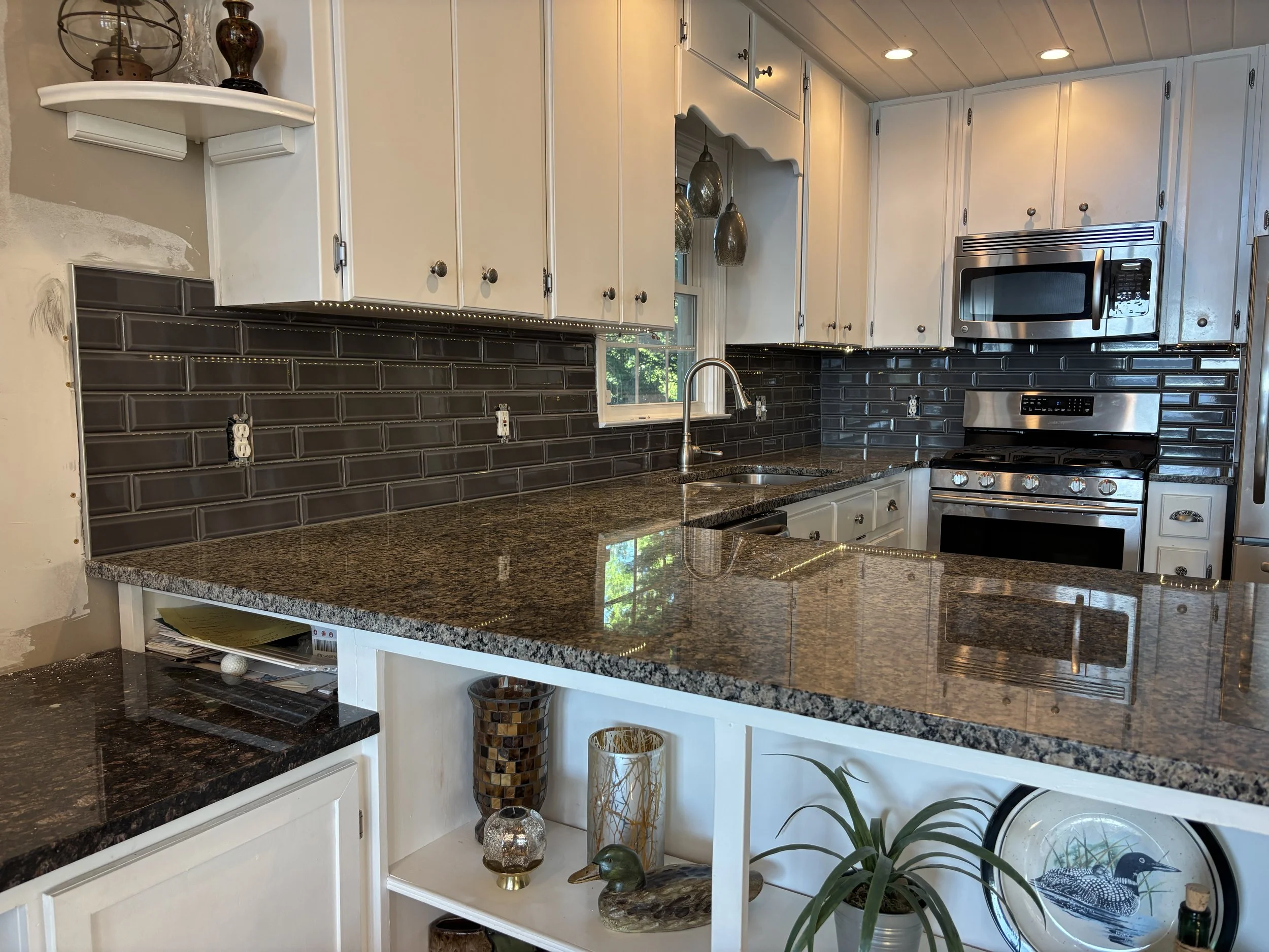 Kitchen with white cabinets, black tile backsplash, stainless steel microwave and oven, granite countertop, window with natural light, decorative vases, plants, and ornaments.