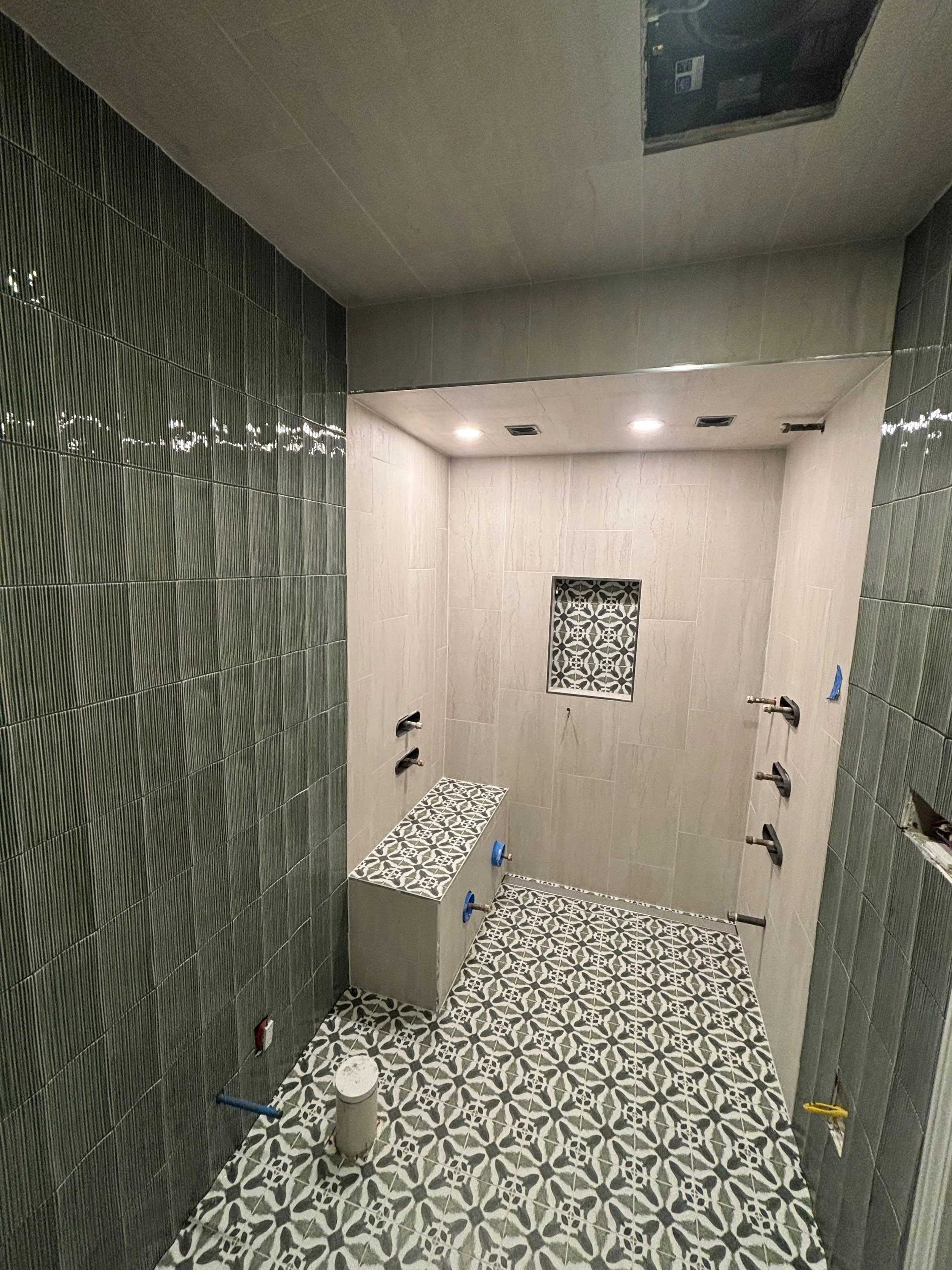 View of a bathroom with green tiled walls, patterned floor tiles, several uninstalled fixtures, and construction materials.