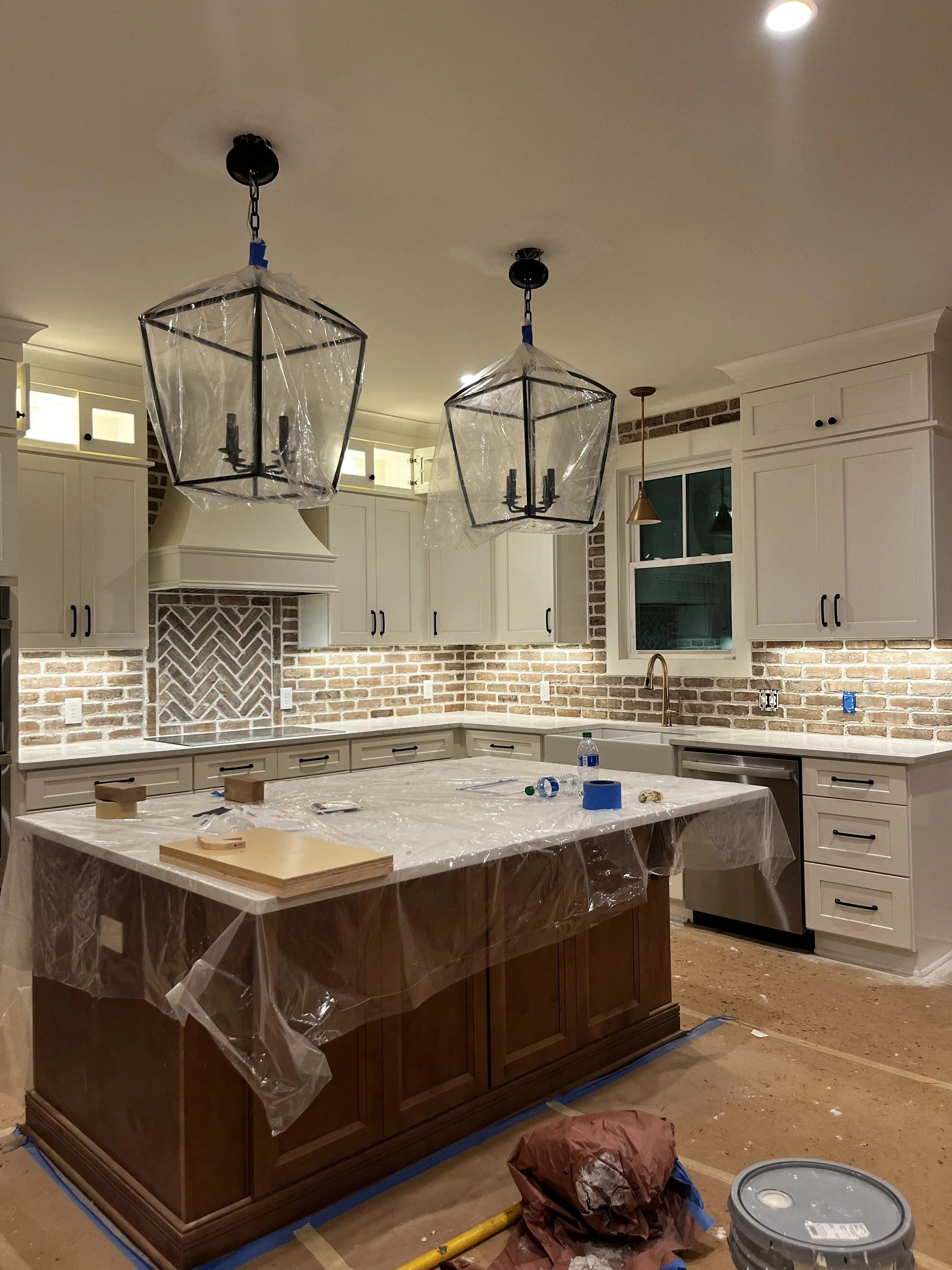 Kitchen under renovation with a brick backsplash, white cabinets, a central island with a dark wood base, and hanging pendant lights covered in plastic. Construction supplies, including a paint bucket, are on the floor.