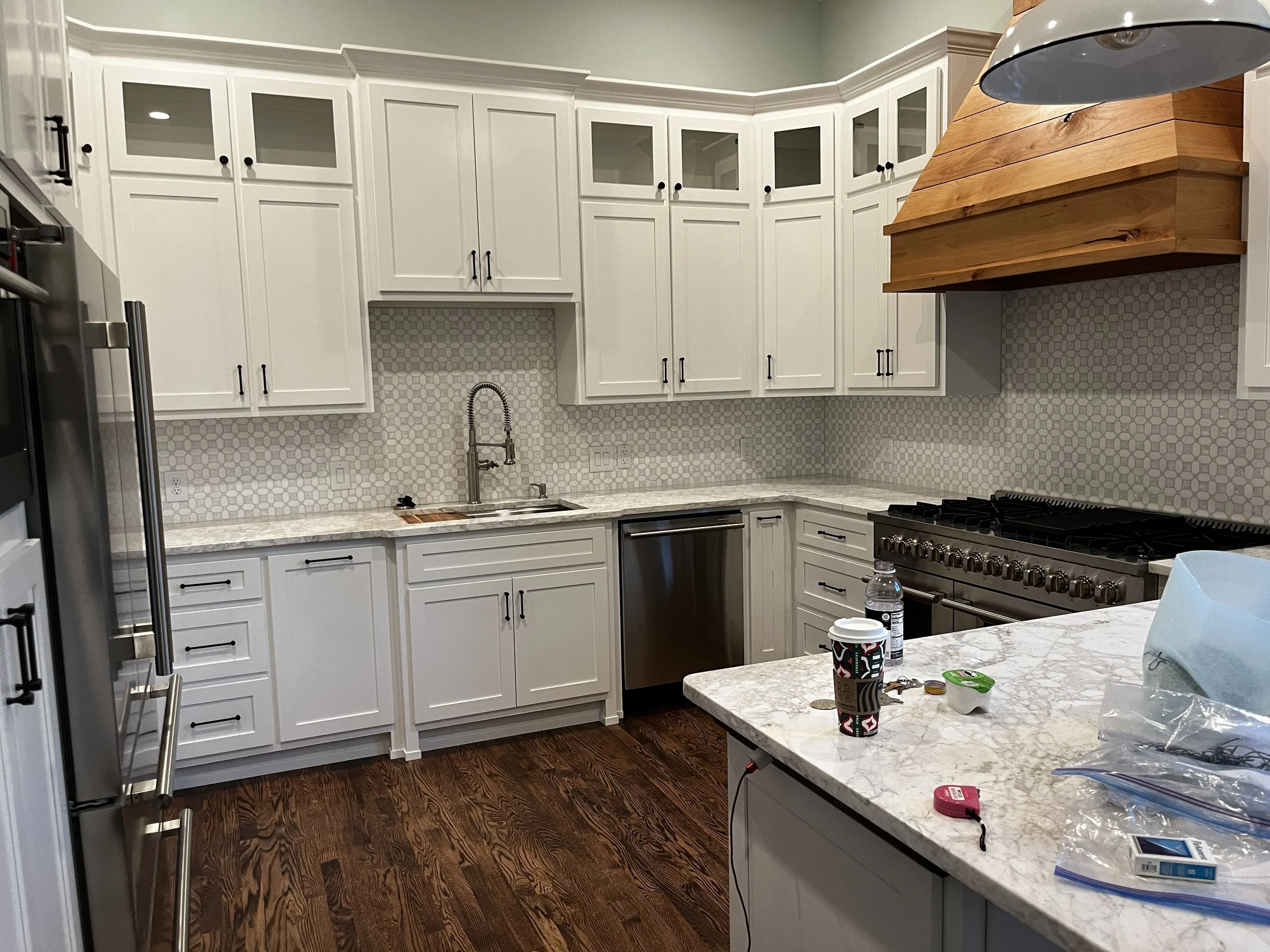 Modern kitchen with white cabinets, granite countertops, stainless steel appliances, and a wooden range hood. Items on the counter include a paper cup, food containers, a water bottle, coins, and small objects.