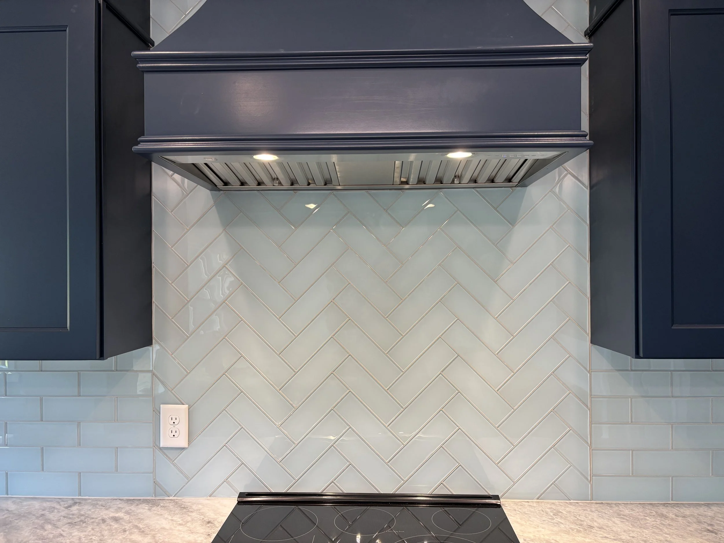 A modern kitchen range hood with built-in lights mounted on a clock wall above a tiled backsplash with diamond-shaped tiles and a double electrical outlet.