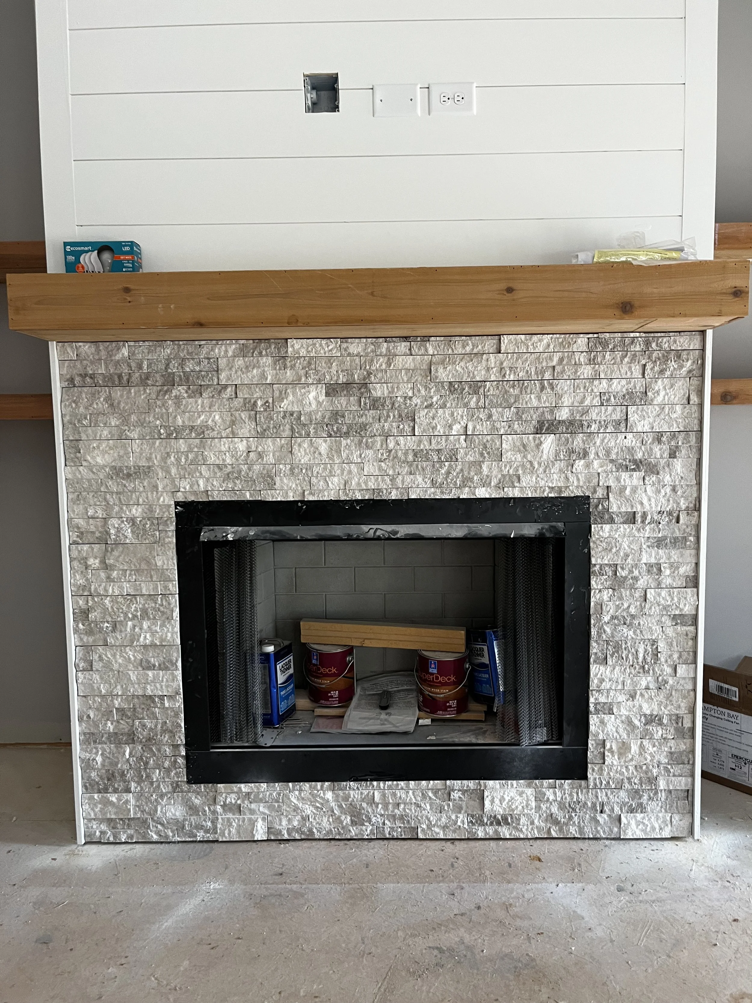 Fireplace with stone surround, black frame, and unfinished interior. Construction supplies inside, including paint cans and a piece of wood. Above the fireplace, a wooden mantel and wiring boxes are visible.