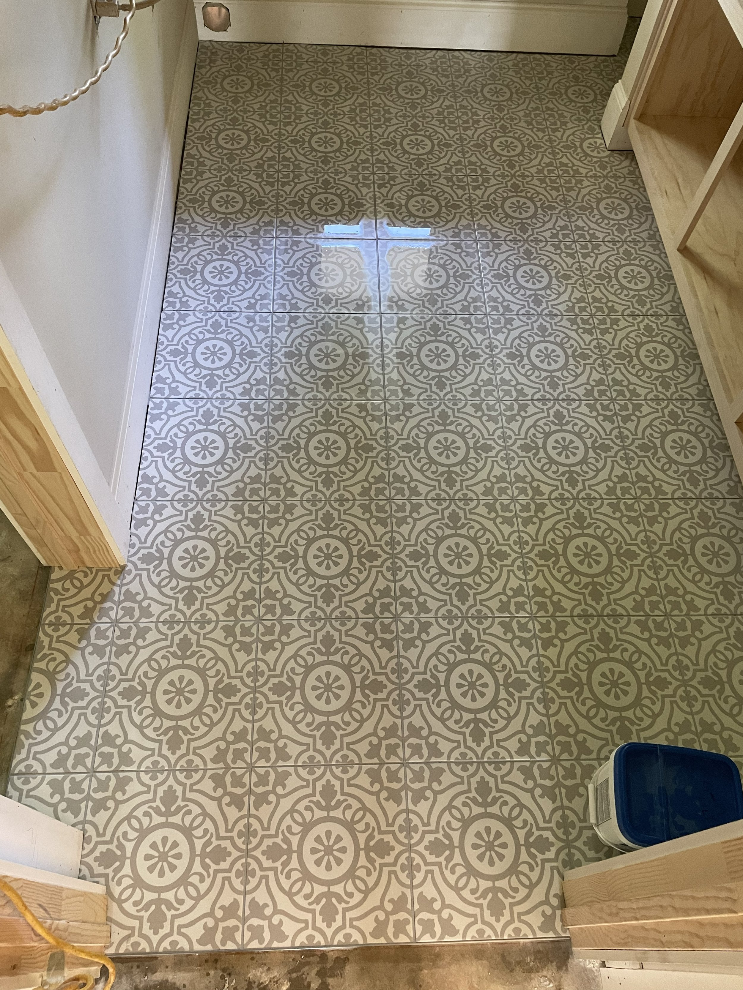 Floor with decorative patterned tiles in gray and white in a room or hallway. Part of a doorway and a wooden shelf are visible.