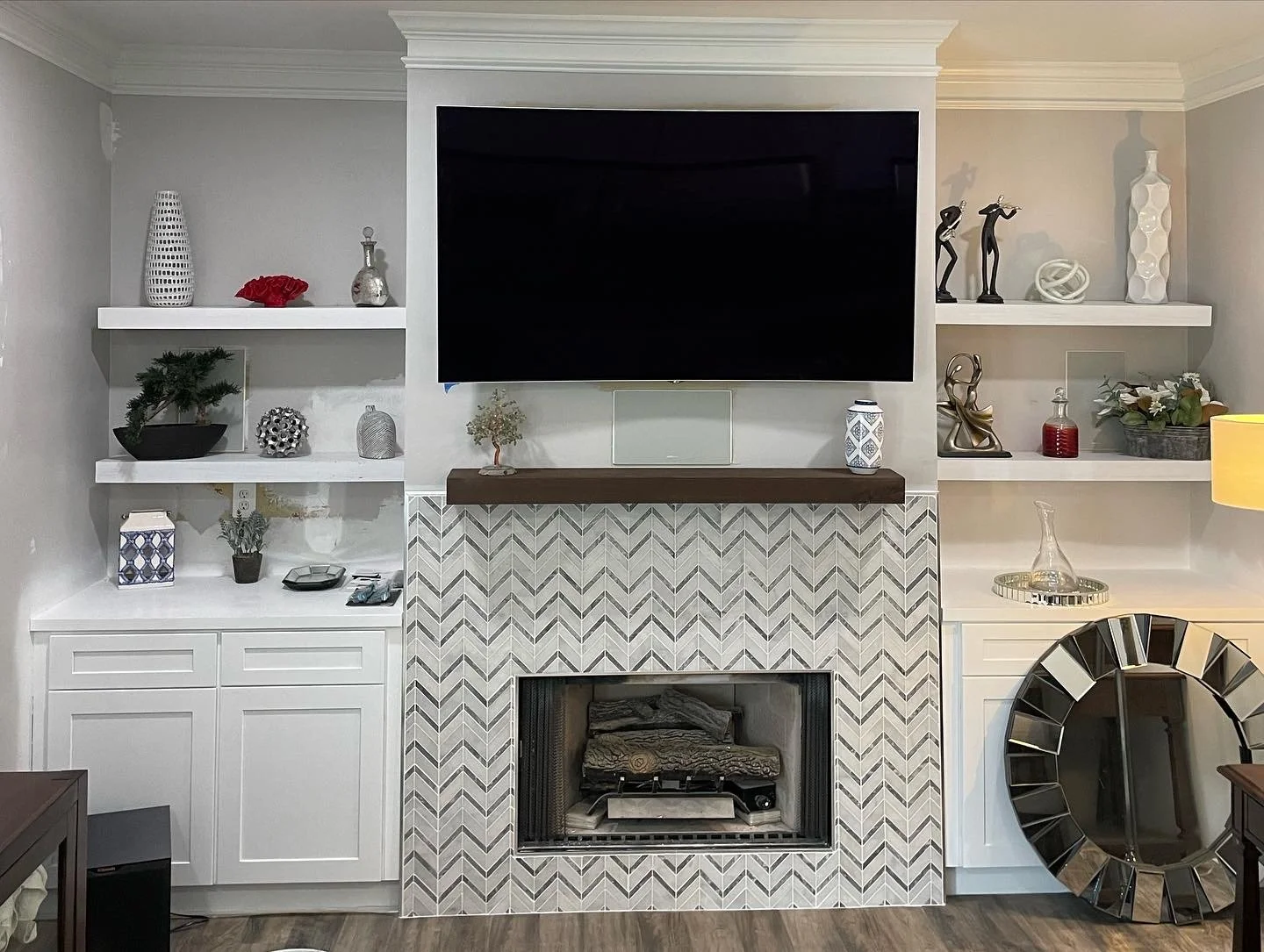 Living room with a fireplace and shelves, wall-mounted flat screen TV, decorative items on shelves including vases, sculptures, a small potted plant, and a mirror with a mirror frame on the right.
