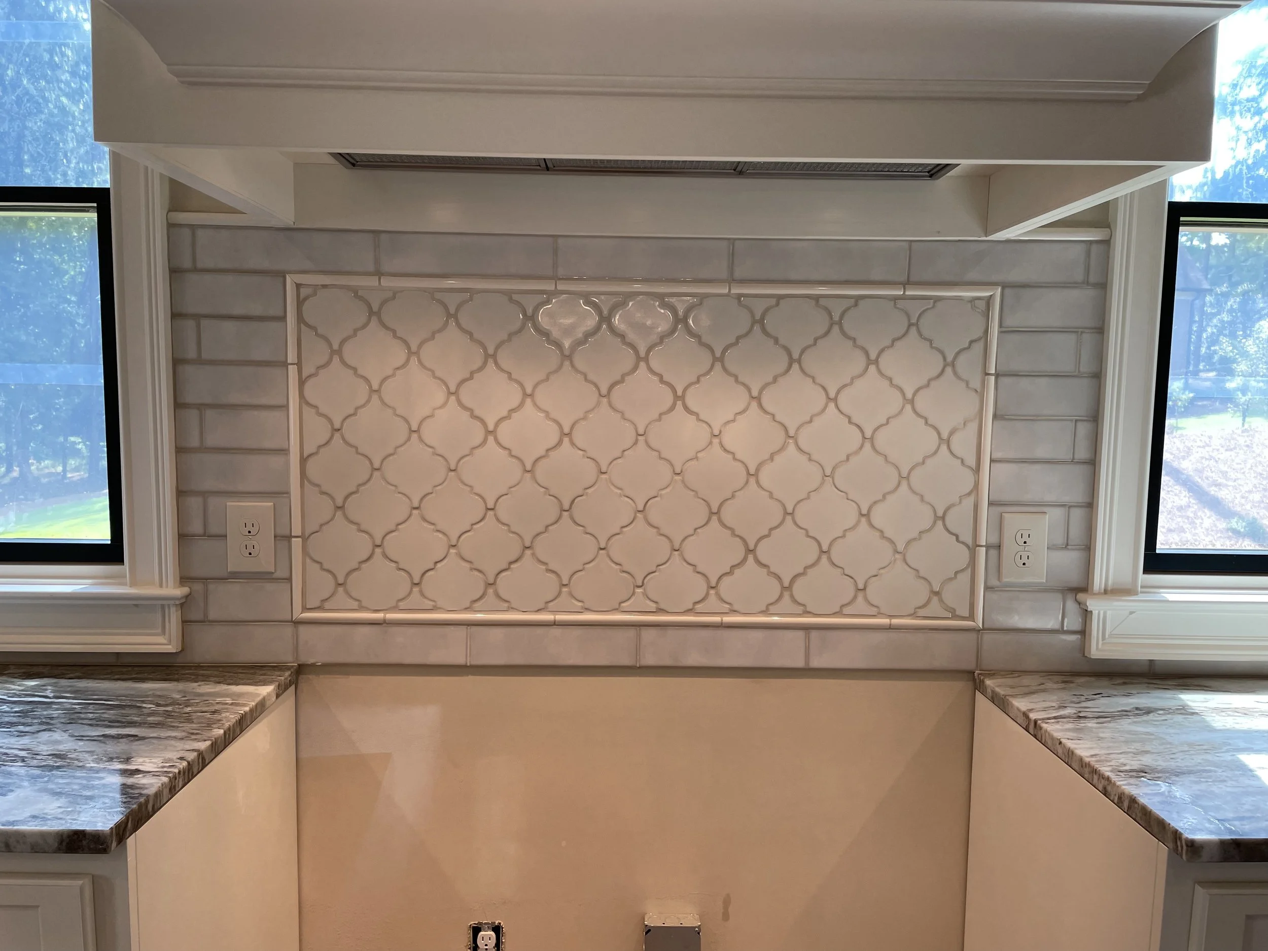 A kitchen backsplash featuring a patterned tile design with a beige and cream color scheme. The backsplash is framed by a white trim and flanked by two windows. Granite countertops are visible at the bottom of the image, and electrical outlets are pr