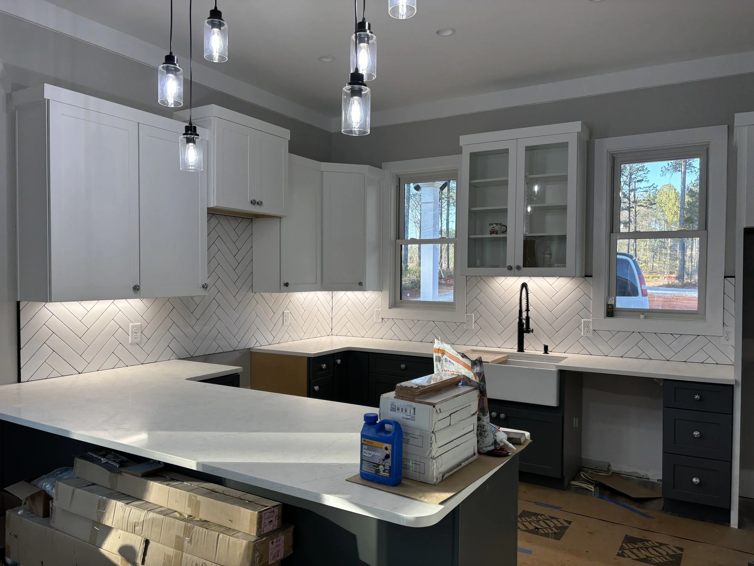 Modern kitchen with white cabinets, black lower cabinets, and a light-colored countertop. Several windows let in natural light, revealing a wooded outdoor scene. Pendant lights hang from the ceiling, and various construction or renovation supplies ar