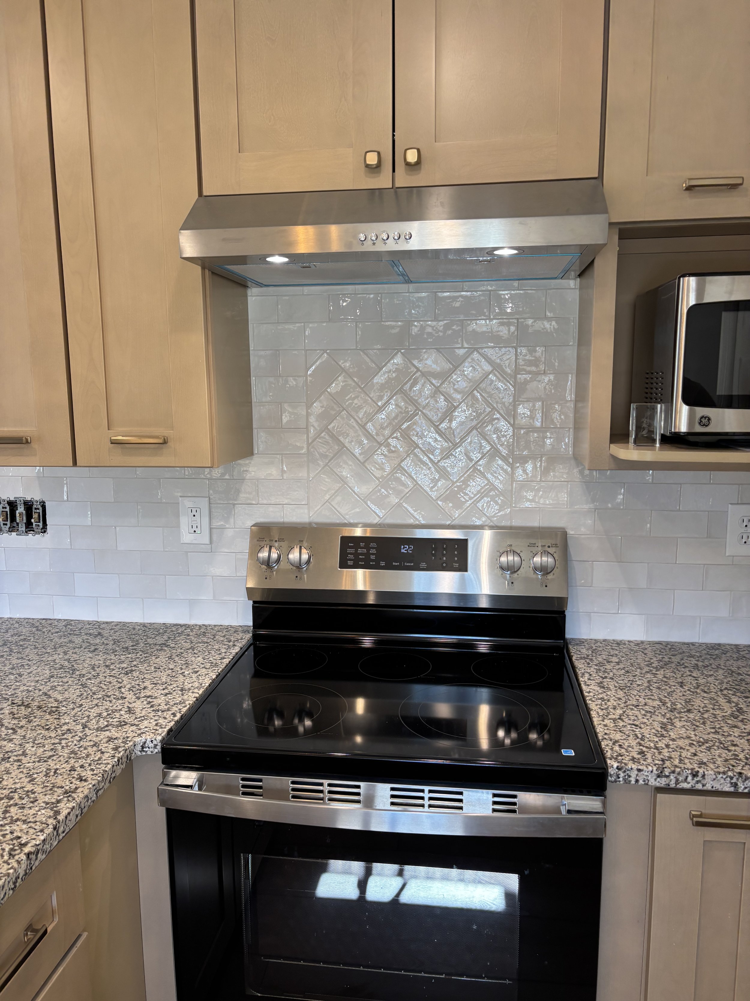 Kitchen stovetop and oven below a stainless steel range hood, with light beige wooden cabinets above and a granite countertop on either side.