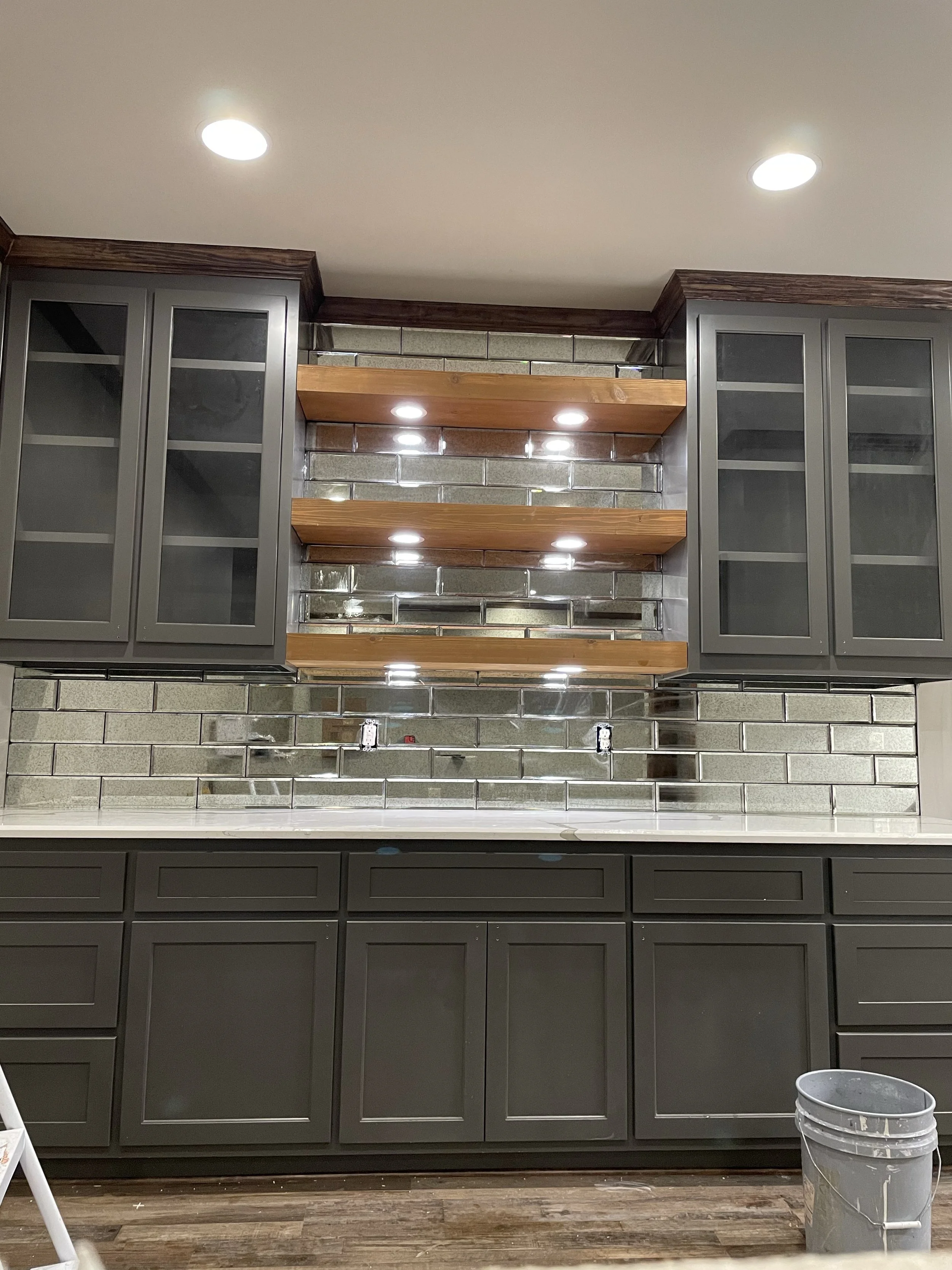 A kitchen wall with grey cabinets and glass doors, a brick backsplash, and three wooden shelves with built-in ceiling lights. There is a white countertop below and a bucket on the floor to the right.