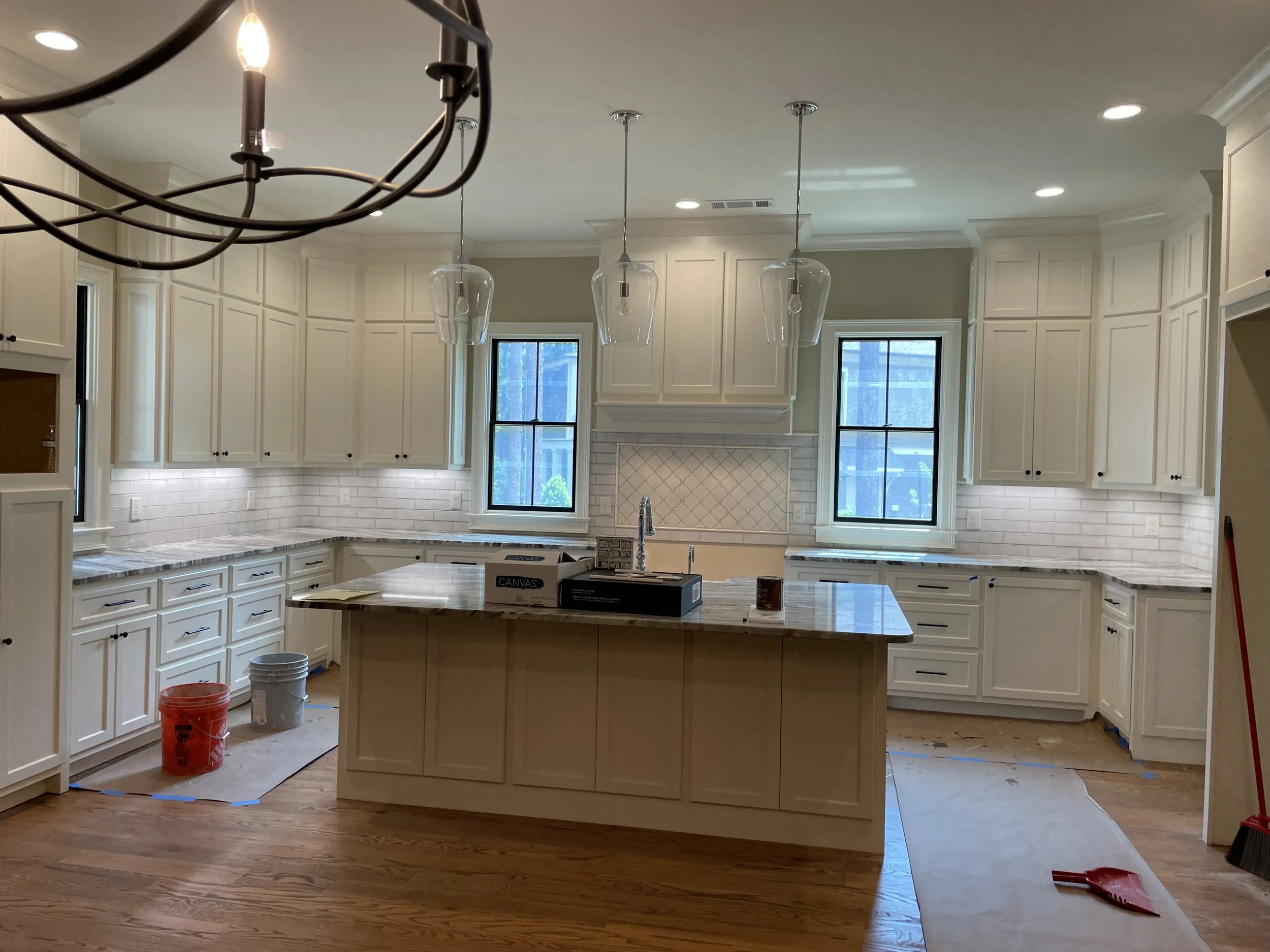 A spacious kitchen under renovation with white cabinets, a center island, pendant lights, and large windows. Construction materials and tools such as buckets, boxes, and cleaning tools are visible.