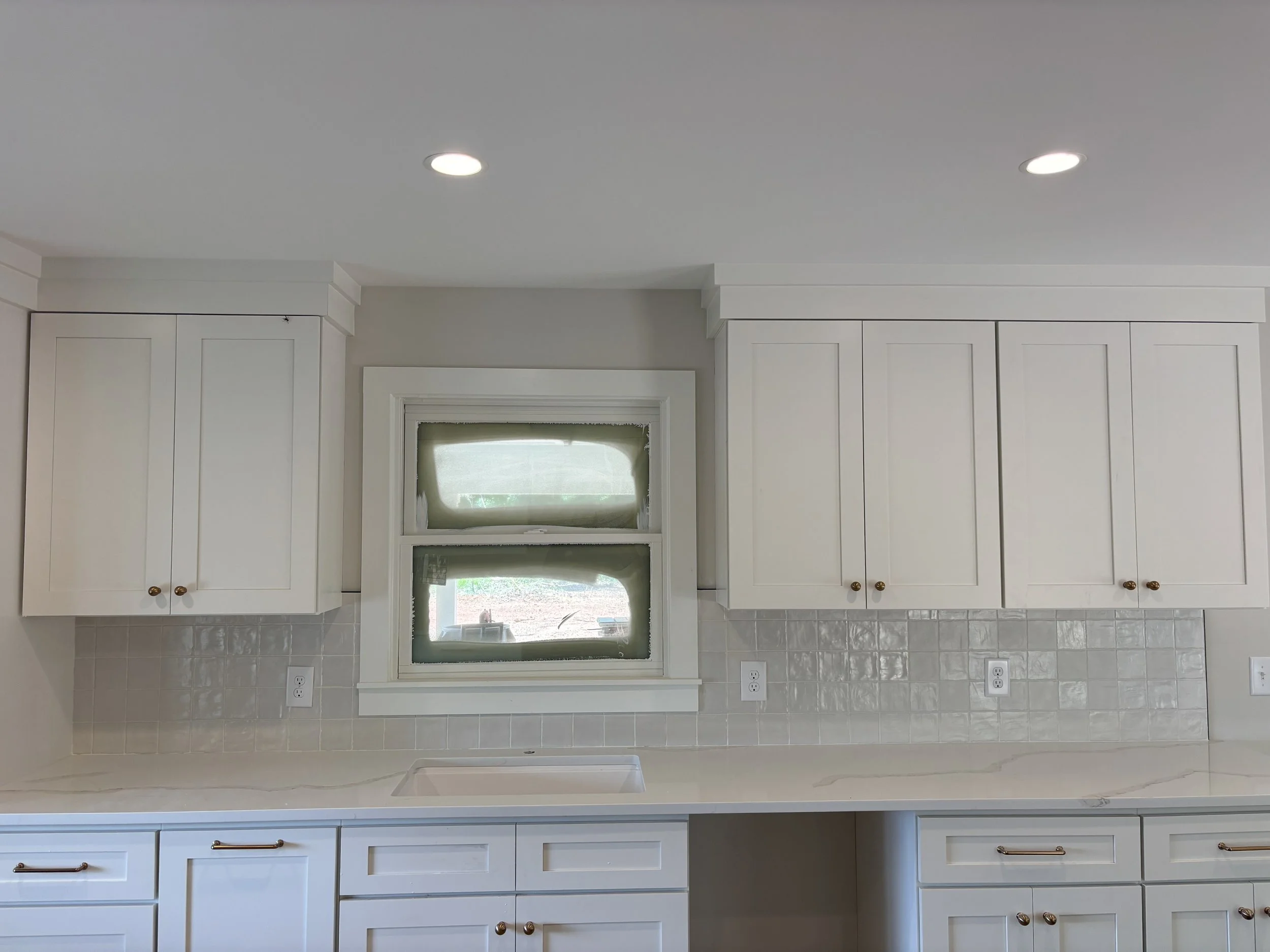 White kitchen cabinets and drawers with gold knobs, a small window with a green blind, and a tiled backsplash. Four electrical outlets are visible on the wall beneath the cabinets.