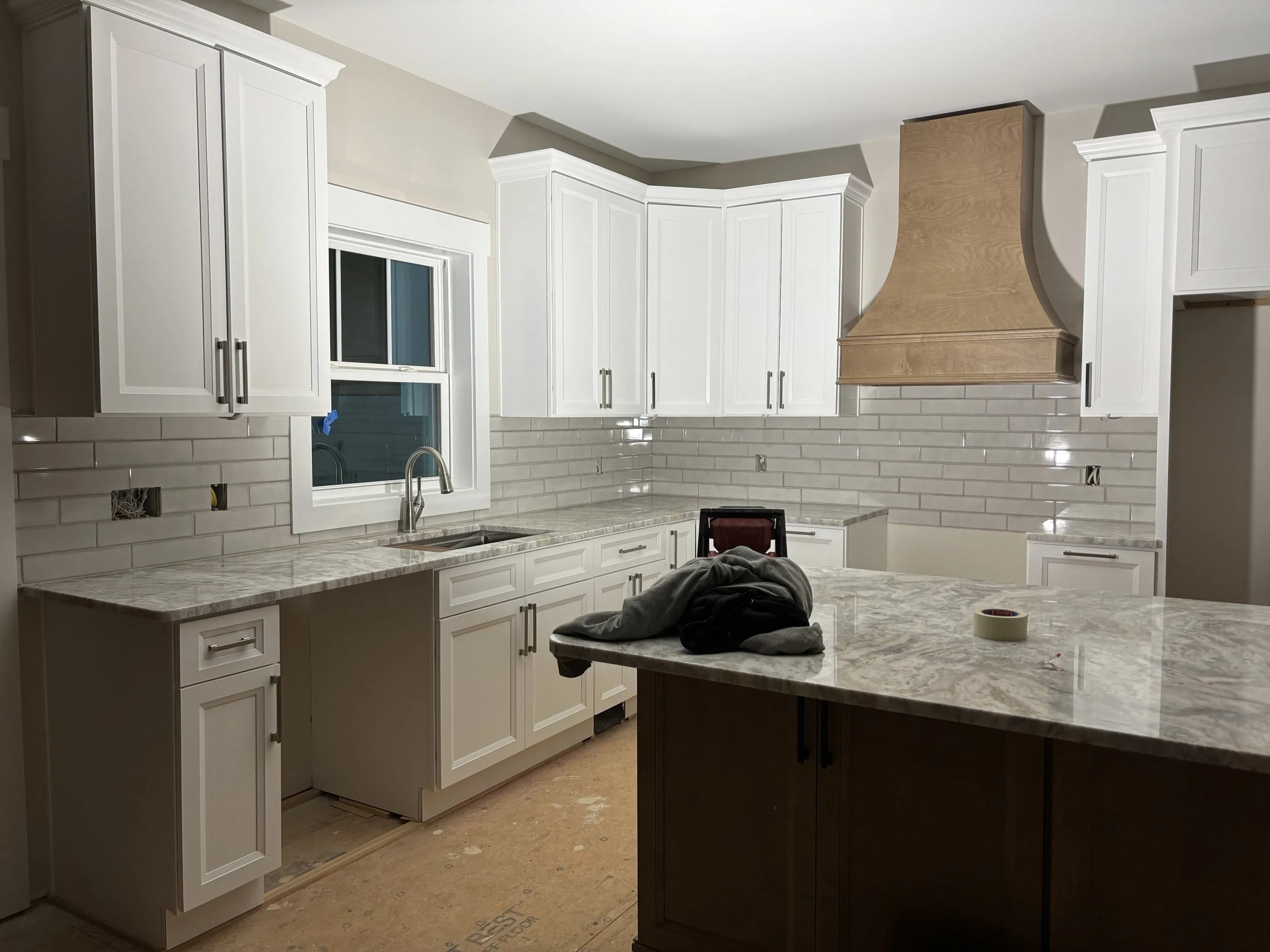 Kitchen with white upper cabinets, a window above the sink, a beige subway tile backsplash, and a brown island with a marble countertop. A wooden range hood is installed over an area for a stove.