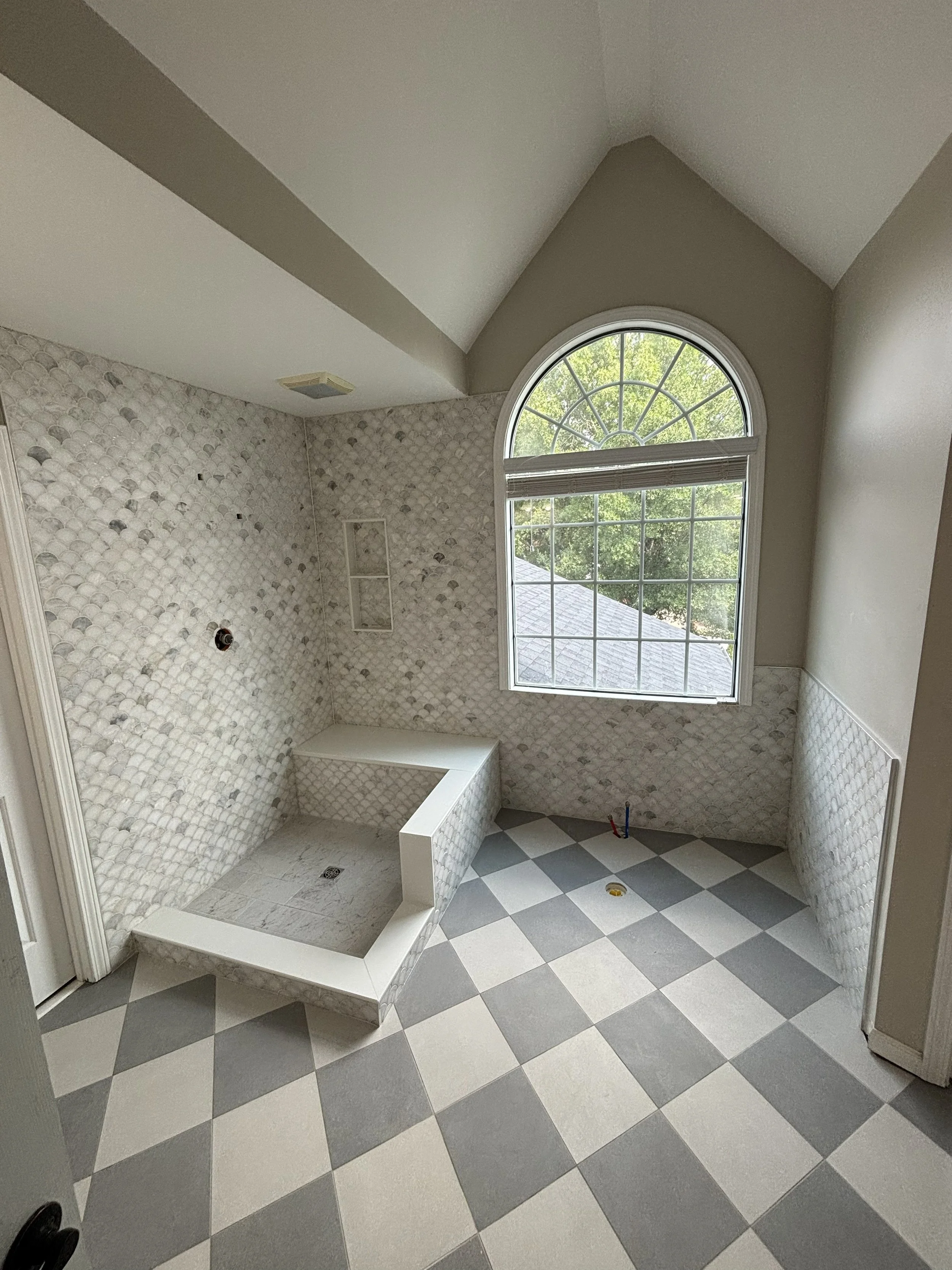 Bathroom with large checkered gray and white floor tiles, a big window with a semi-circular top, and a shower area with small pebble-style tiles, unfinished with plumbing fixtures and some construction materials.