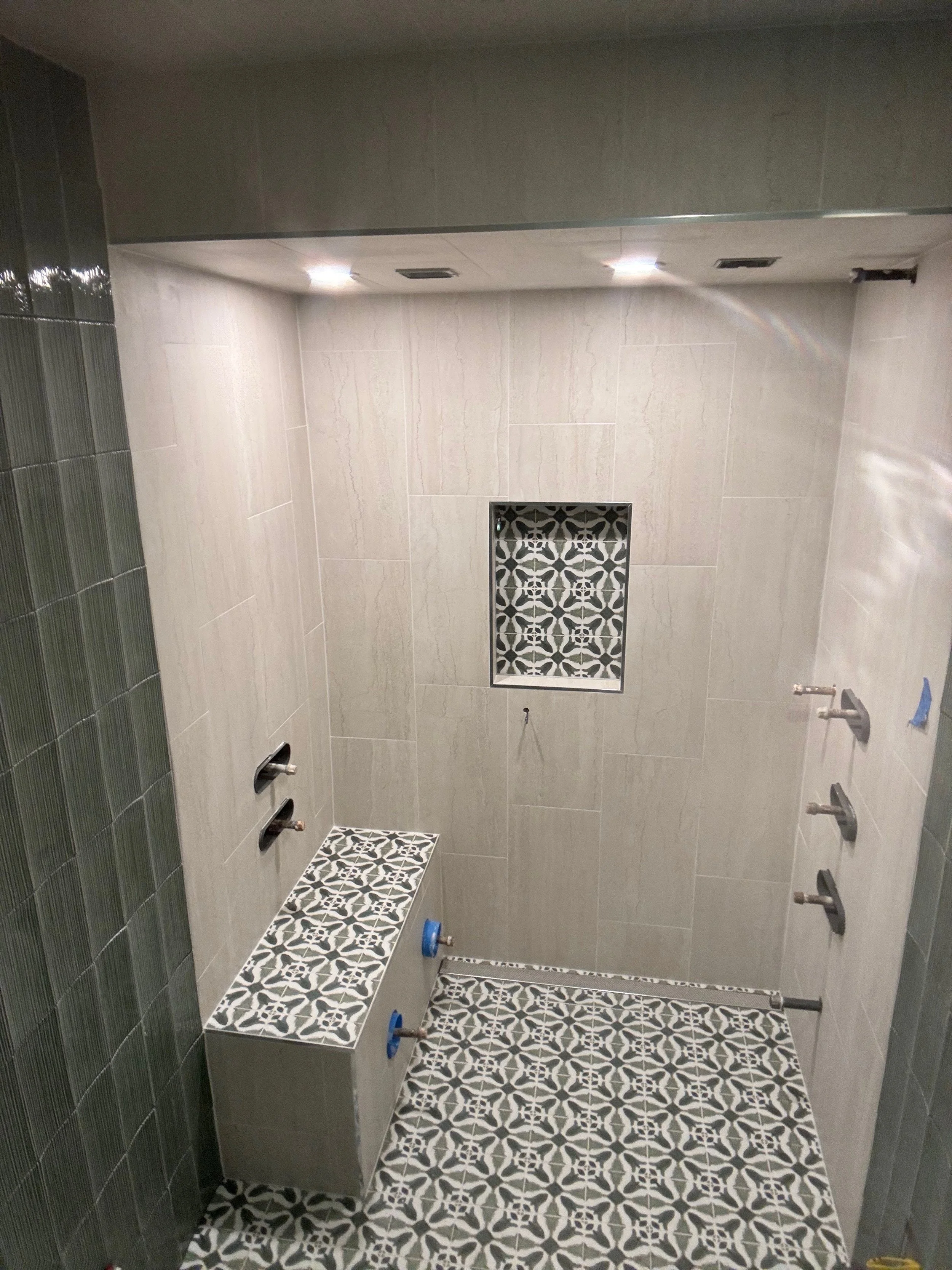 This is a photo of a tiled shower area that is under construction or renovation. The shower has light-colored large wall tiles, patterned floor tiles, and a decorative niches with black and white geometric tiles. The shower fixtures are not installed