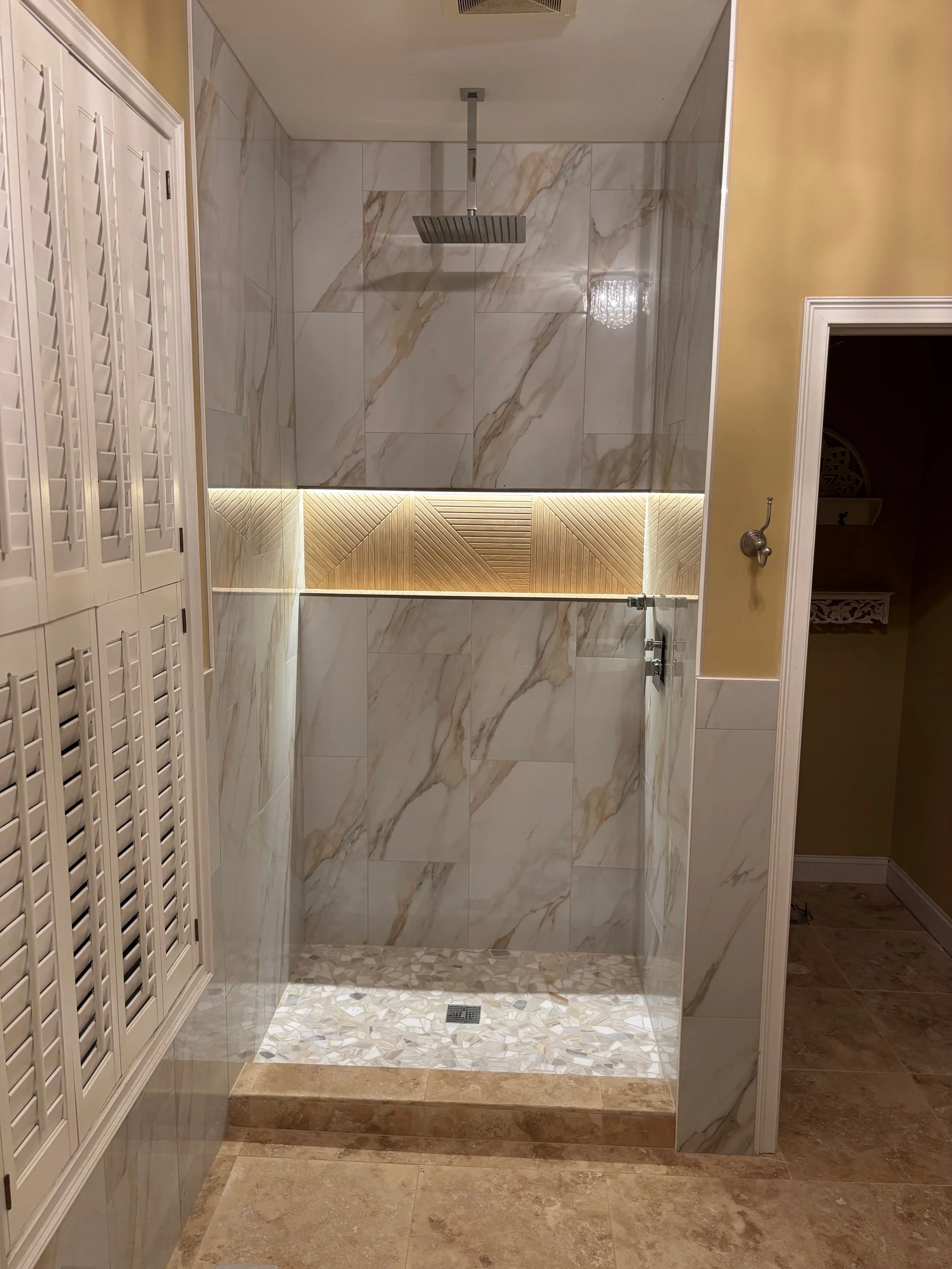 A modern walk-in shower with large marble tiles, a rainfall showerhead, under-lighting design accents, and a stone border at the entrance, adjacent to a beige and white decorative wall and a small alcove.