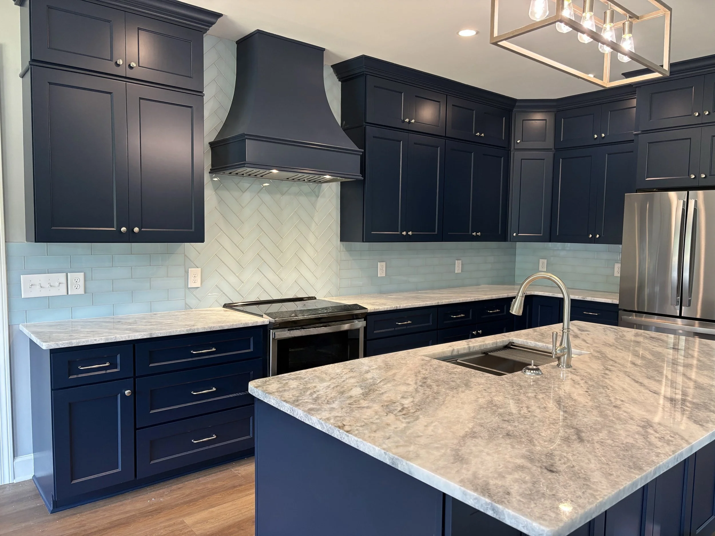 Modern kitchen with dark blue cabinets, white marble countertops, light blue backsplash with a herringbone pattern, stainless steel appliances including a refrigerator, and a rectangular chandelier with multiple bulbs hanging from the ceiling.