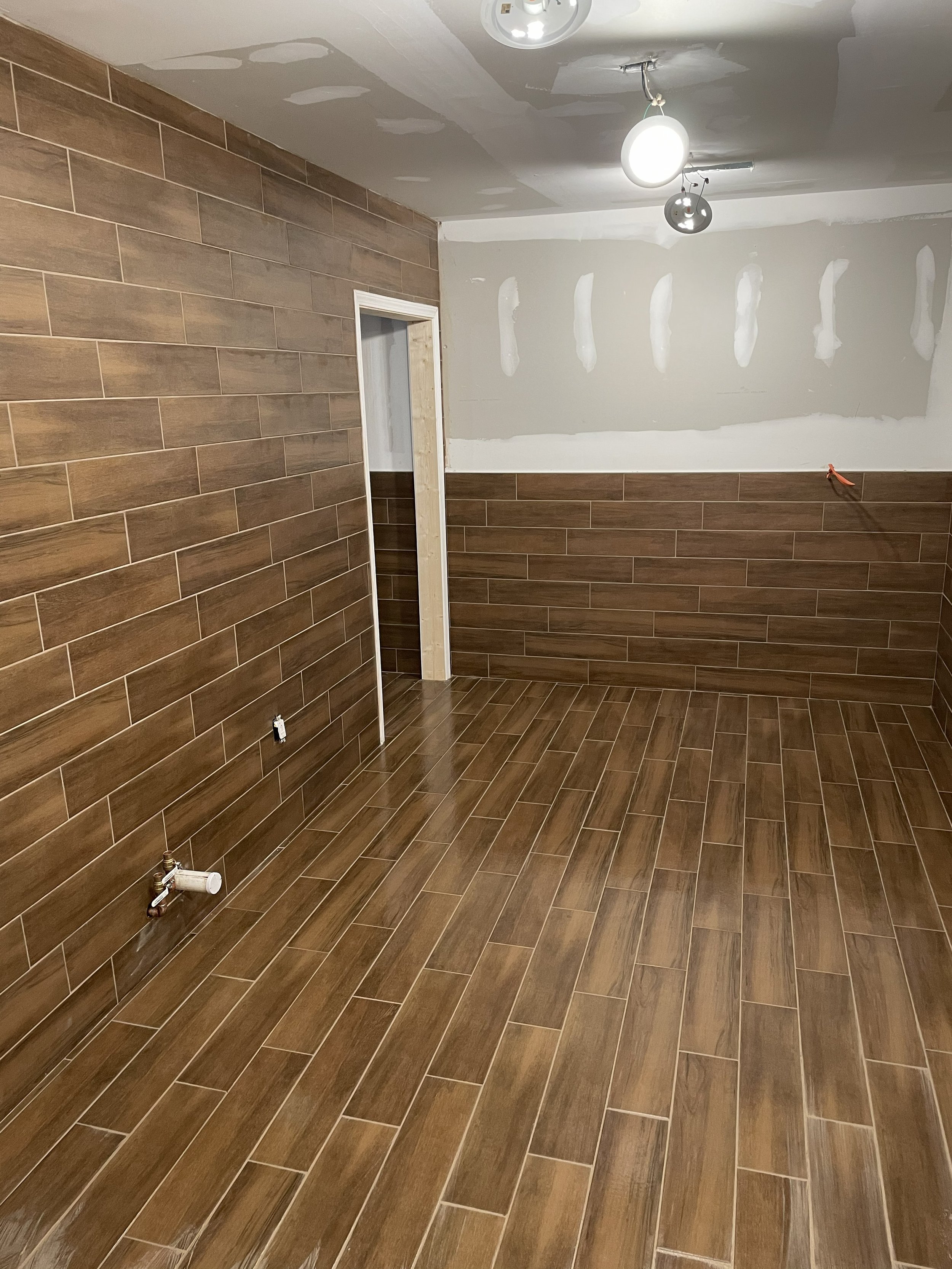 Room under renovation with brown tiled floor and half-tiled walls, unfinished drywall, and exposed light fixtures.