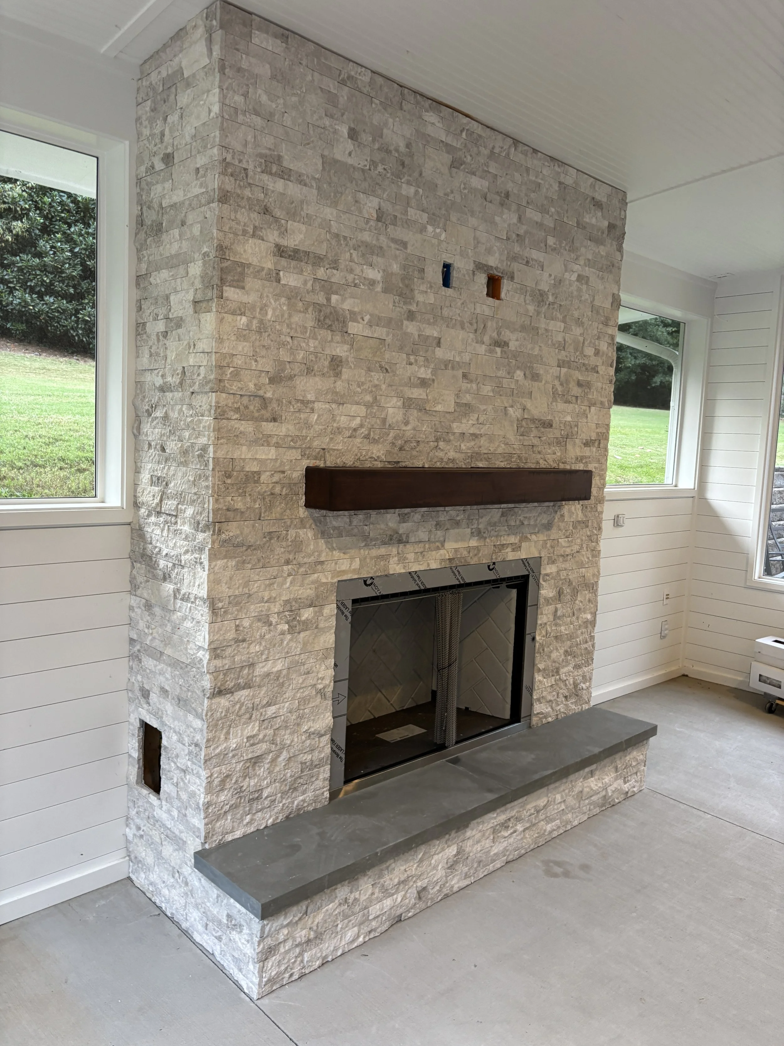 Interior view of a fireplace with stone veneer, unfinished interior, and white siding walls, with windows showing a grassy outdoor area.