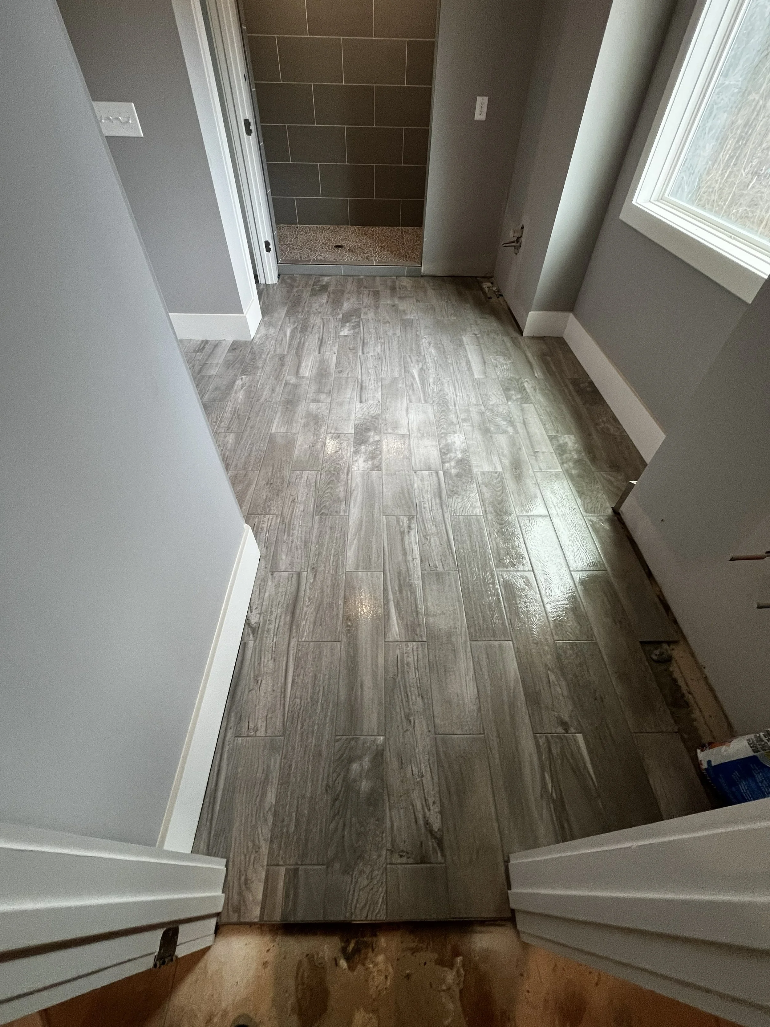 Interior view of a room with gray wooden flooring, gray walls, white baseboards, and a doorway leading to a tiled shower area with dark tiles. A window with white trim is visible on the right side.