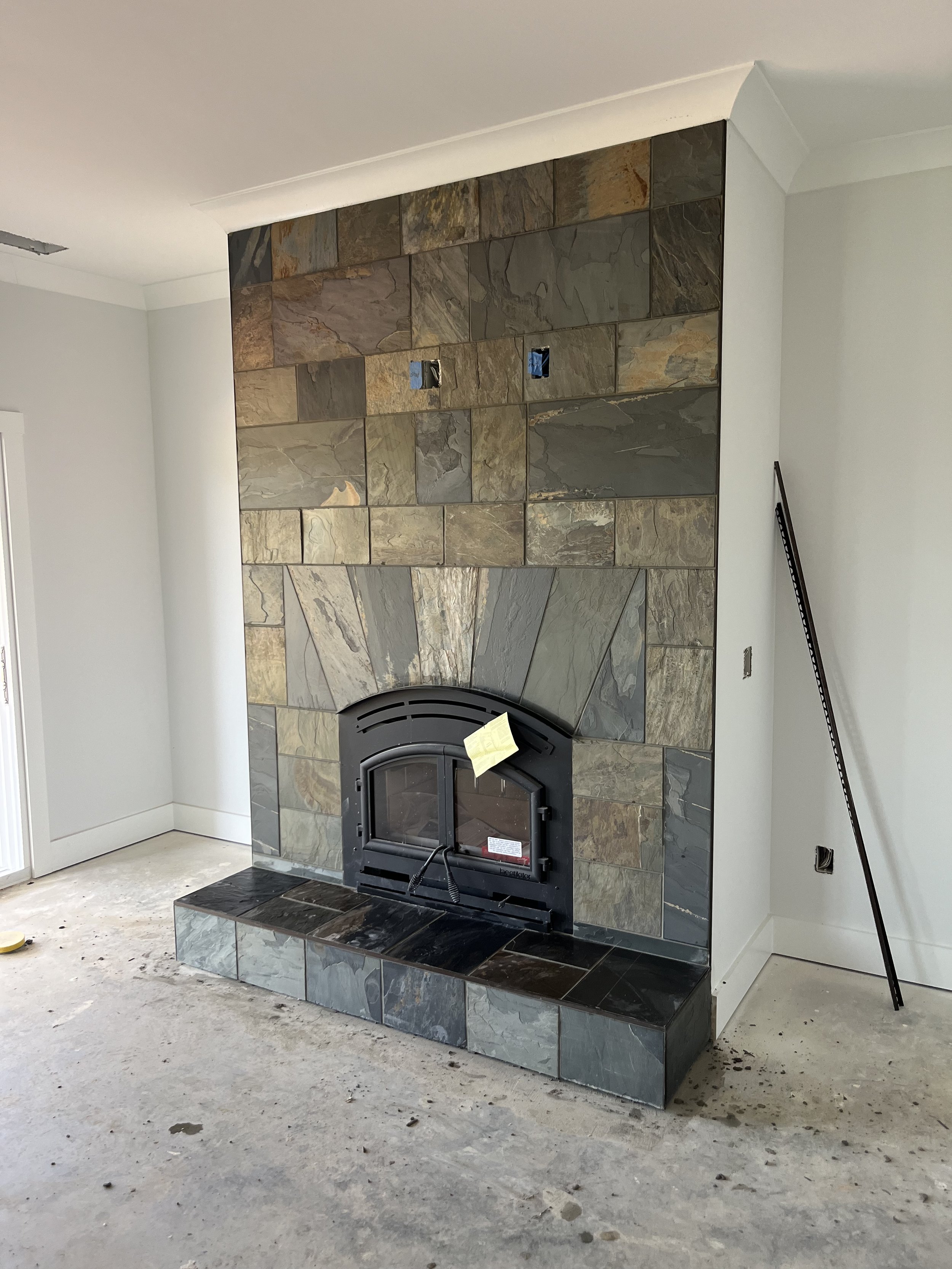 A partially finished living room with a slate tile fireplace. The fireplace has a black metal insert and a stone hearth, with some electrical outlets and a strip of blue painter's tape. The floor is unfinished concrete with some debris, and the walls