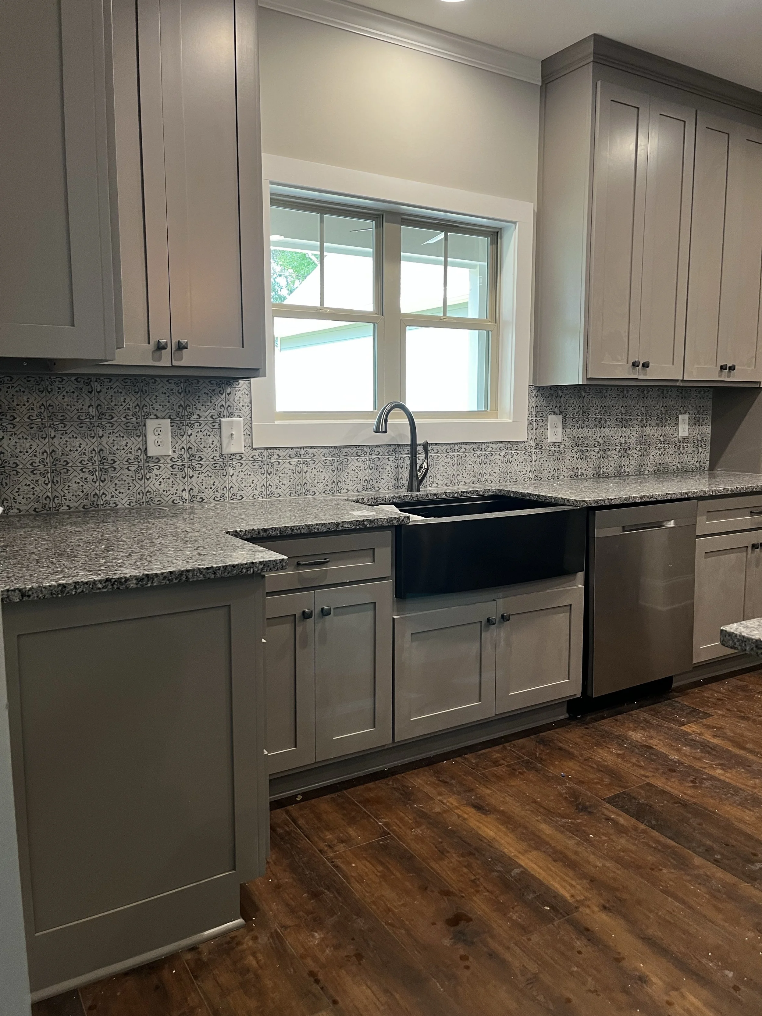 A kitchen with gray cabinets, granite countertops, a window above the sink, a backsplash with patterned tiles, and hardwood flooring.