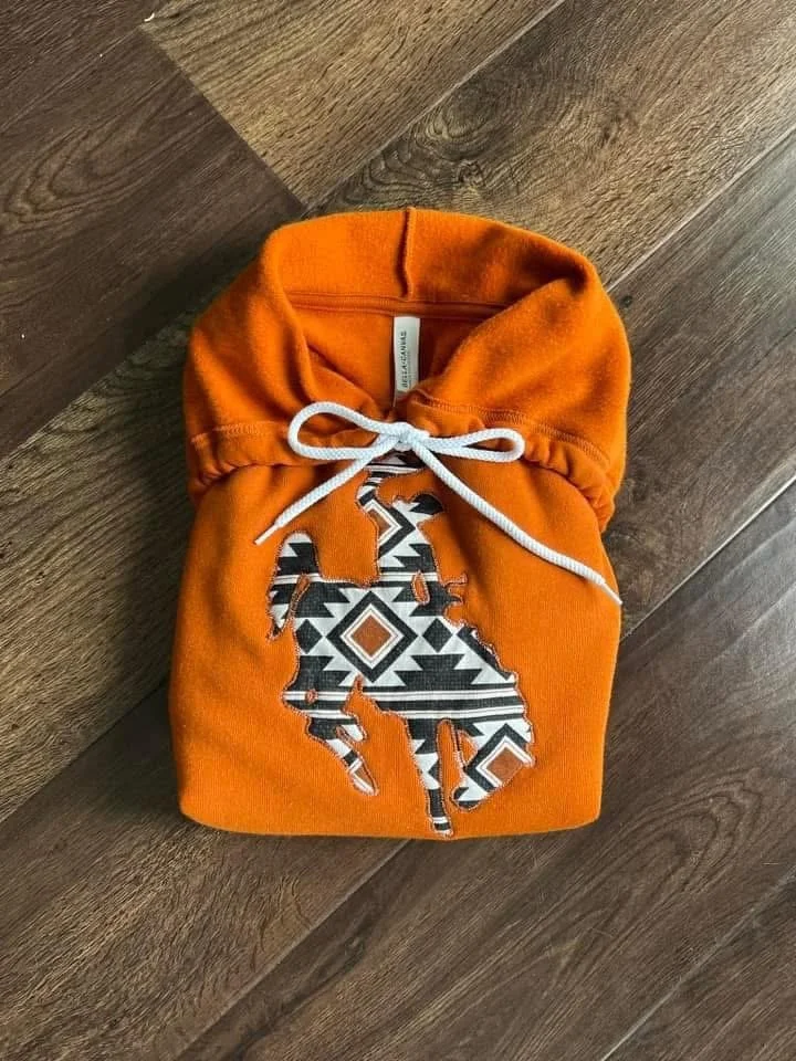 Orange hoodie with a white drawstring, featuring a bucking horse silhouette with a black and white tribal pattern on a wooden floor.