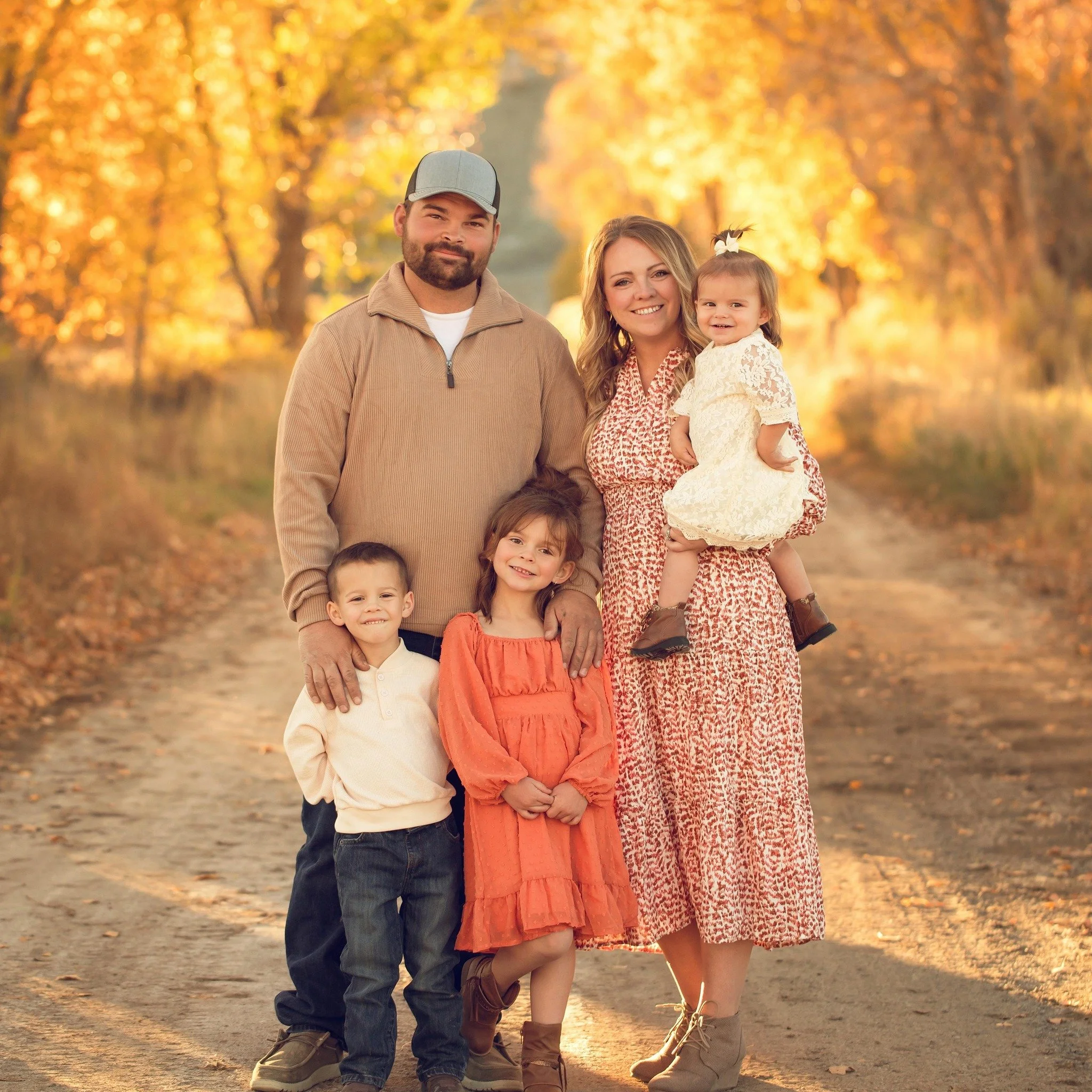 Family of six standing on a dirt path surrounded by autumn trees with golden leaves, smiling at the camera.