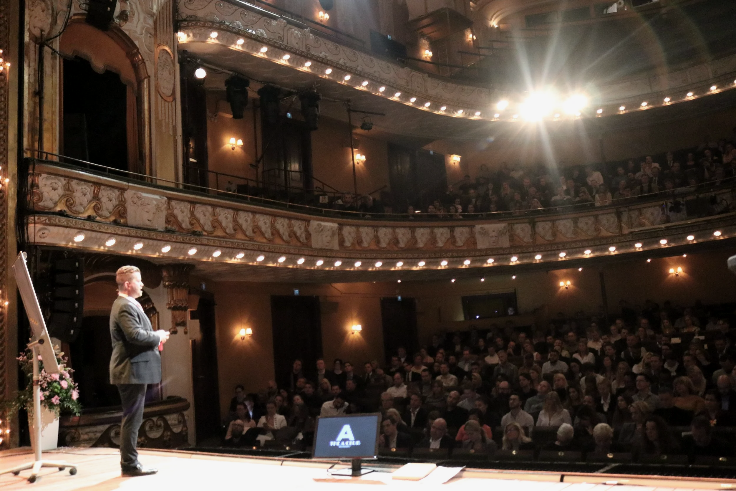 A man in a suit giving a presentation on a stage in a large ornate theater with a balcony, audience members seated and listening, bright stage lighting.